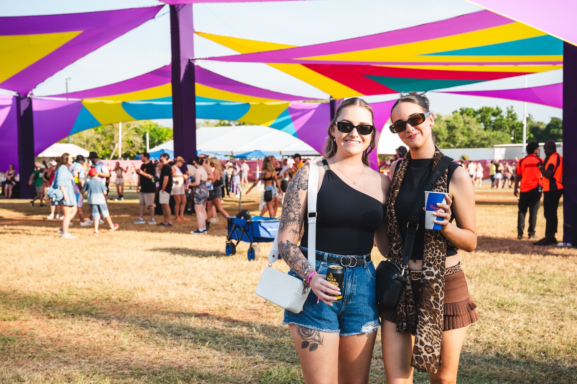 Two women in sunglasses smiling under colorful canopy at an outdoor festival, holding drinks. People and trees in the background.