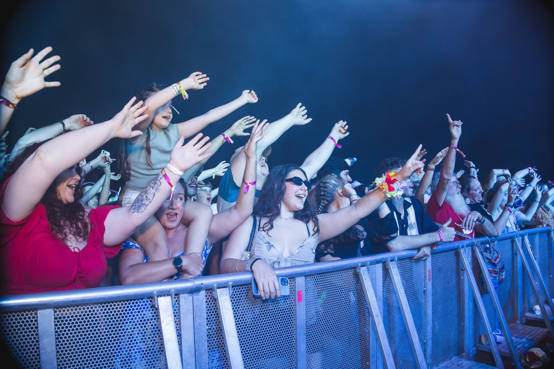 A lively crowd at a concert, with people raising their hands and cheering near a barrier under vibrant stage lighting.