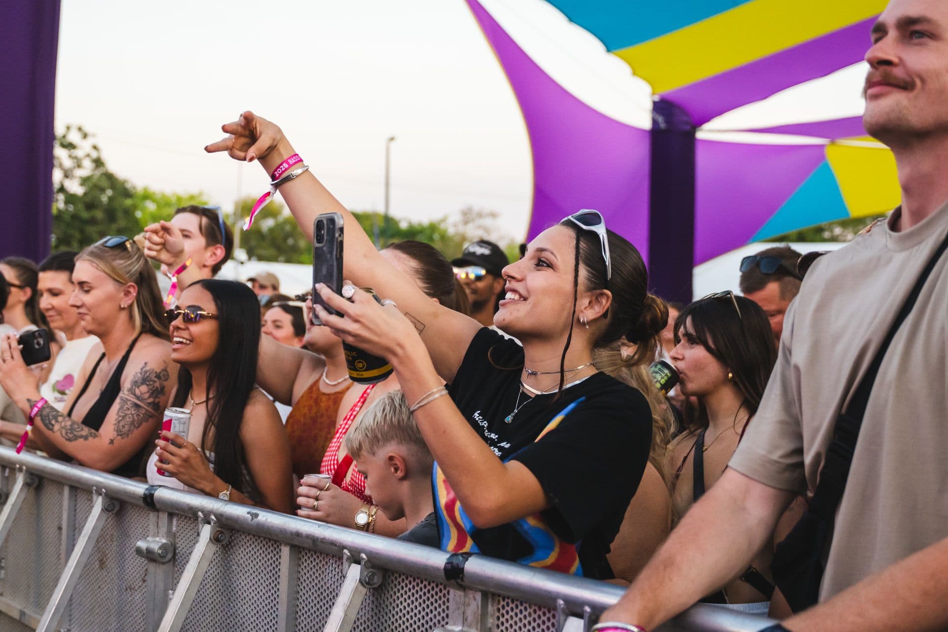 People enjoying a lively outdoor concert, some holding phones to capture the moment, under colourful canopy structures.