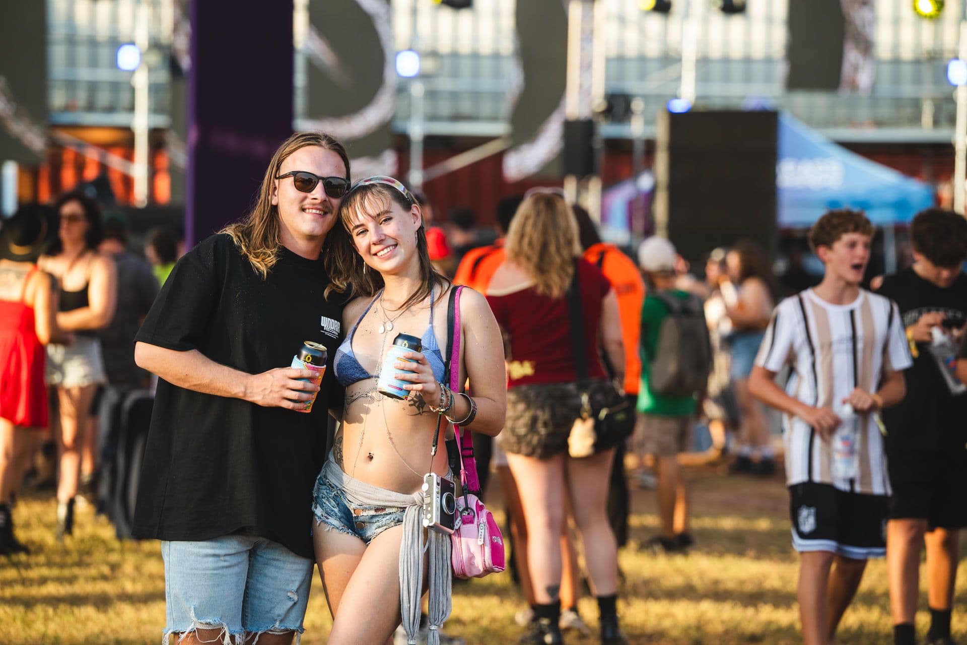 A smiling couple poses at an outdoor festival, holding drinks. People and colorful tents are visible in the sunny background.