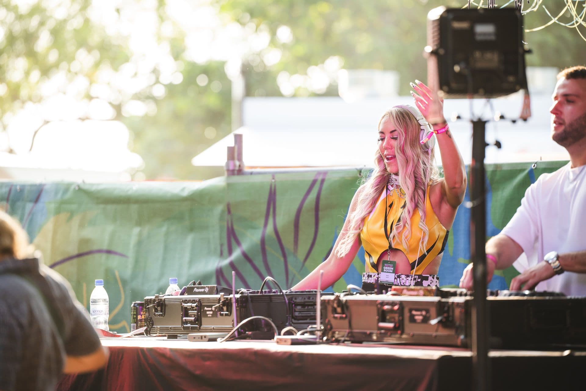 DJ performing at an outdoor event, wearing headphones and a yellow outfit, with turntables and speakers in front, surrounded by greenery and sunlight.
