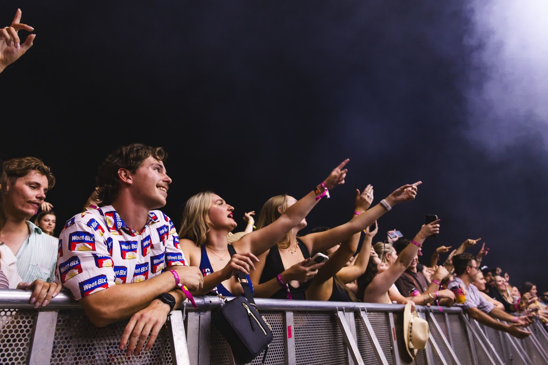 Concertgoers excitedly reach toward the stage, some holding phones. A man in a patterned shirt leans on a barrier, smiling.