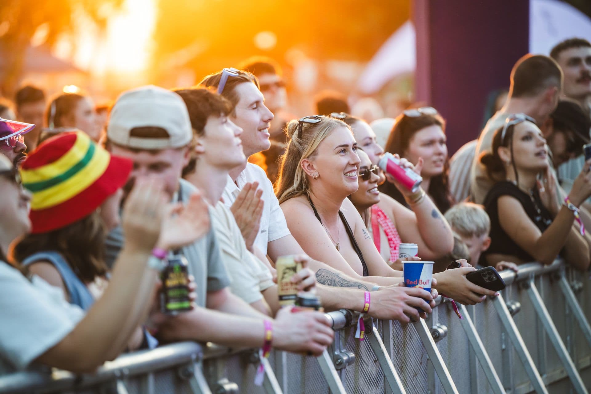 Crowd of people at an outdoor concert, smiling and enjoying the music near a barrier. Sunlight creates a warm, vibrant atmosphere.