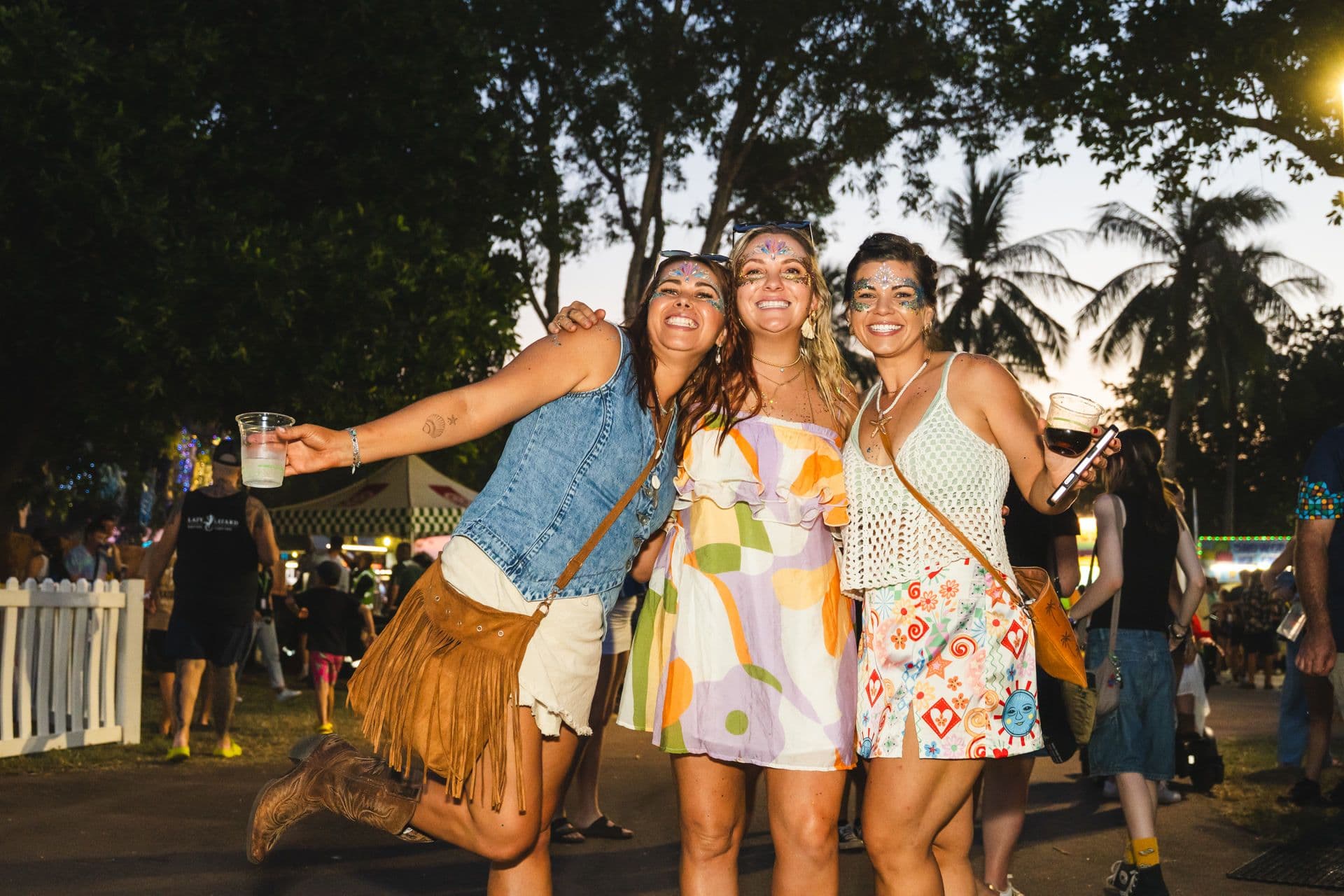 Three women smiling and posing at an outdoor festival, wearing colorful outfits and face paint, with trees and a crowd in the background.