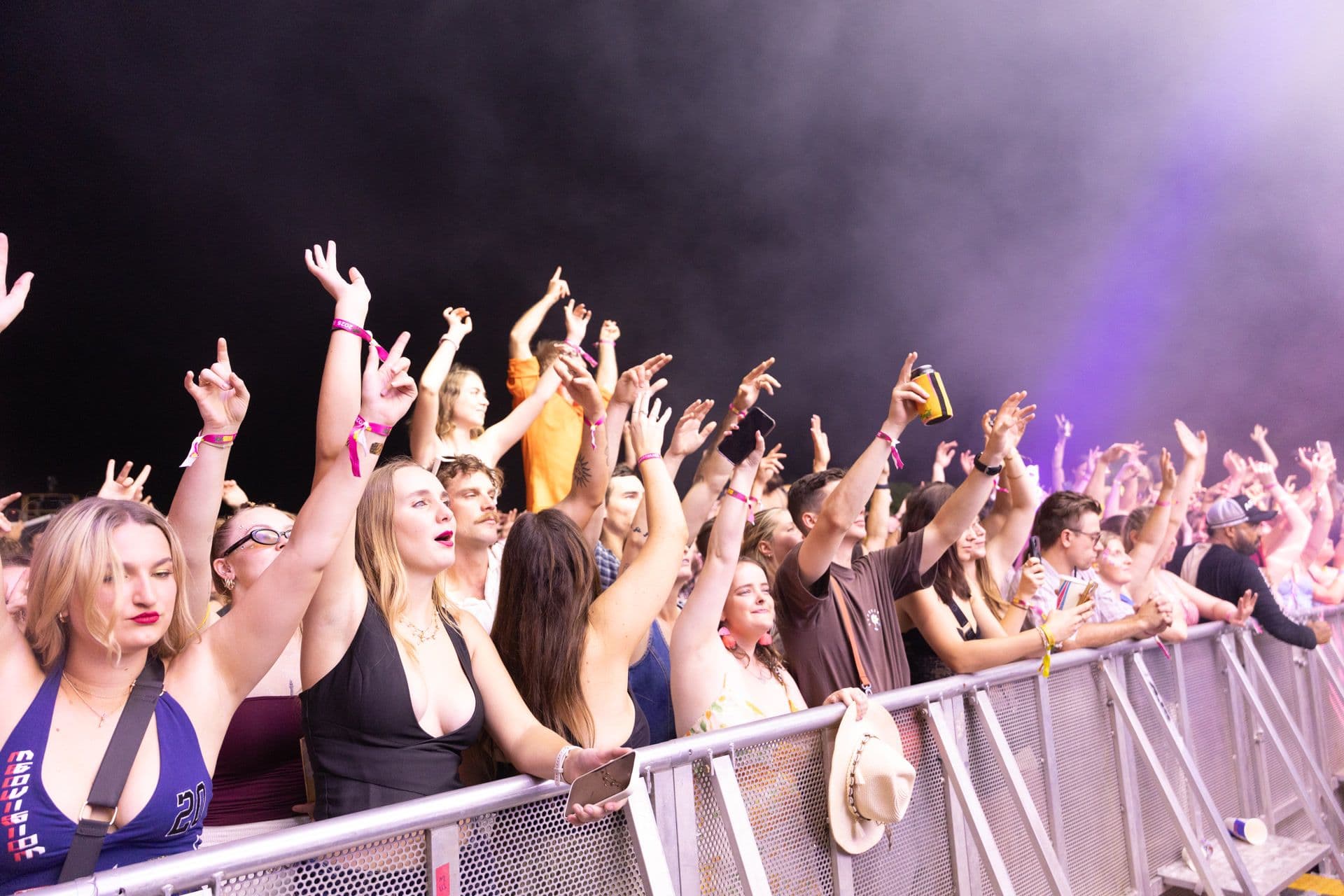 Concert crowd at a barrier, people with raised hands and smartphones, vibrant atmosphere, colorful lighting, and a smoky background.