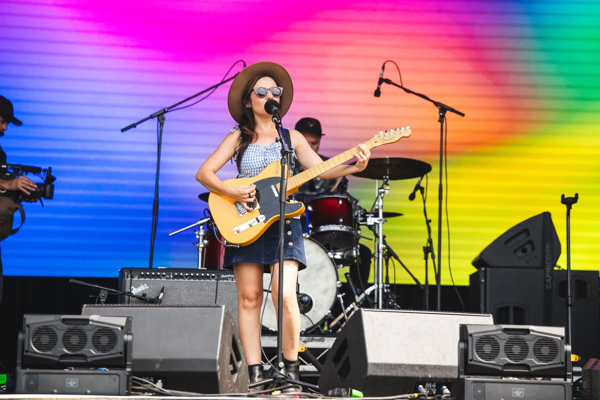 Musician in a hat and sunglasses plays electric guitar on stage with colorful background, drums, and microphones.