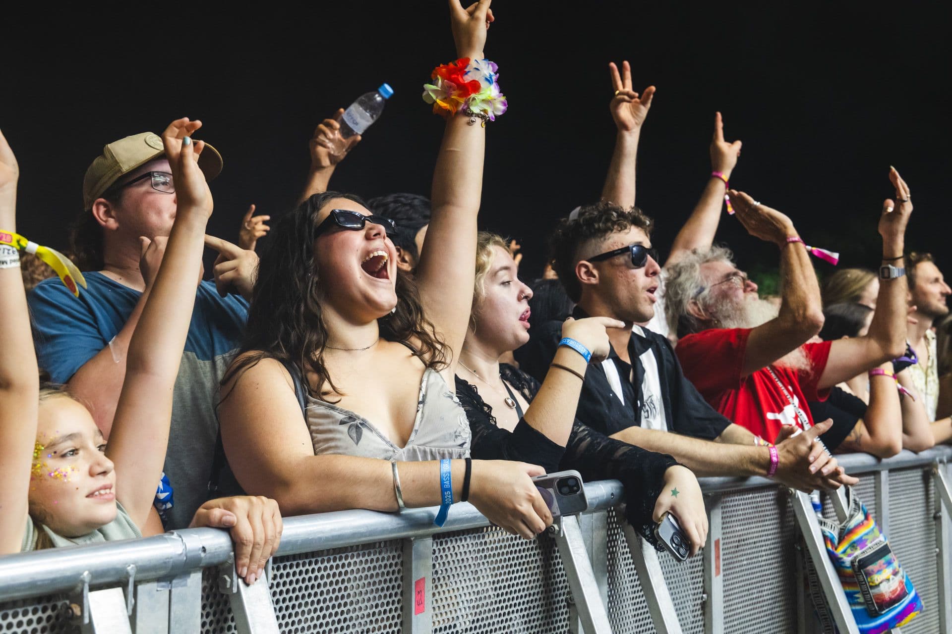 Enthusiastic crowd at a concert, cheering and raising hands, with people wearing sunglasses and colorful accessories near a barricade.