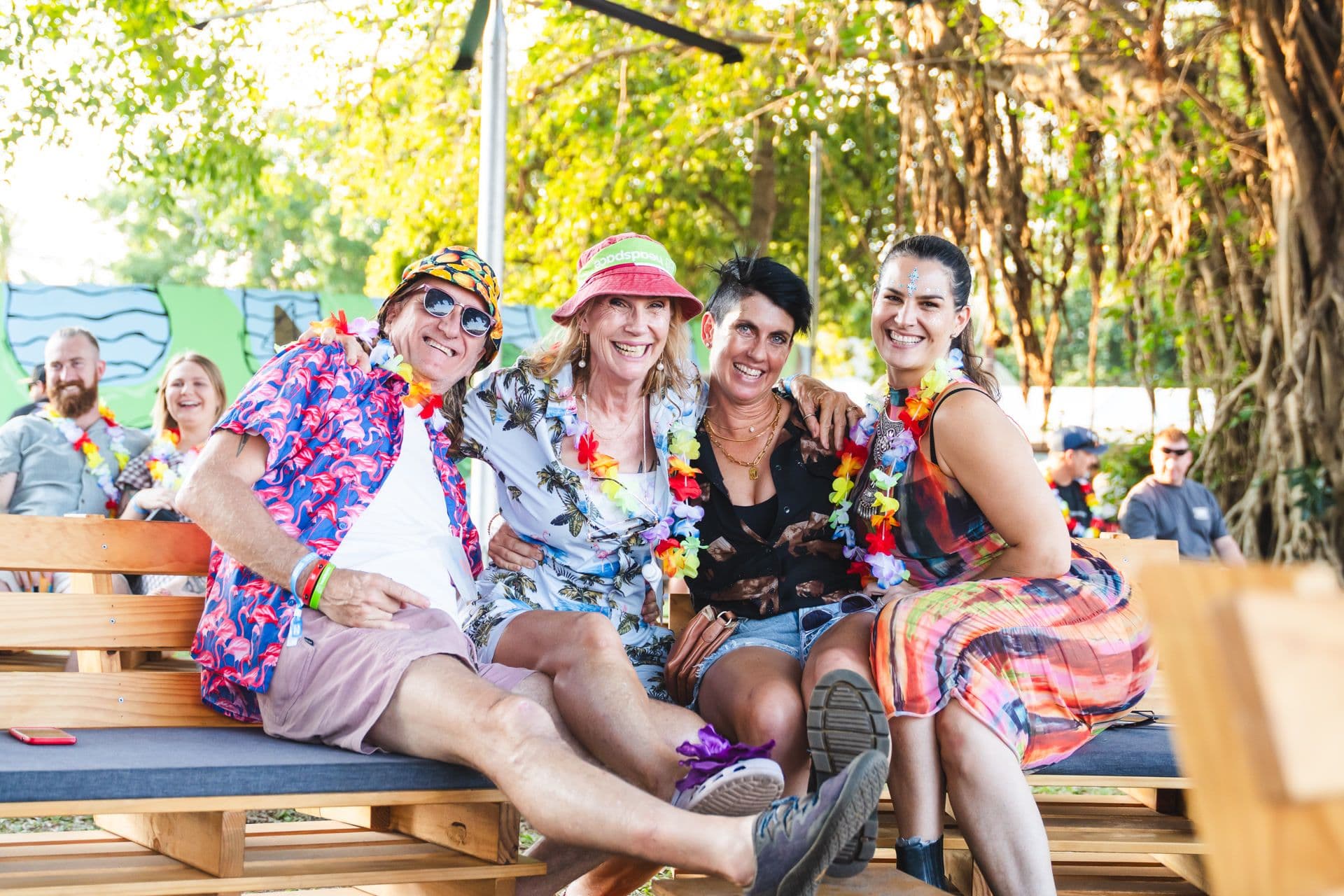 Four people sitting on benches outdoors, wearing colorful clothing and leis, smiling and enjoying a sunny day under trees.