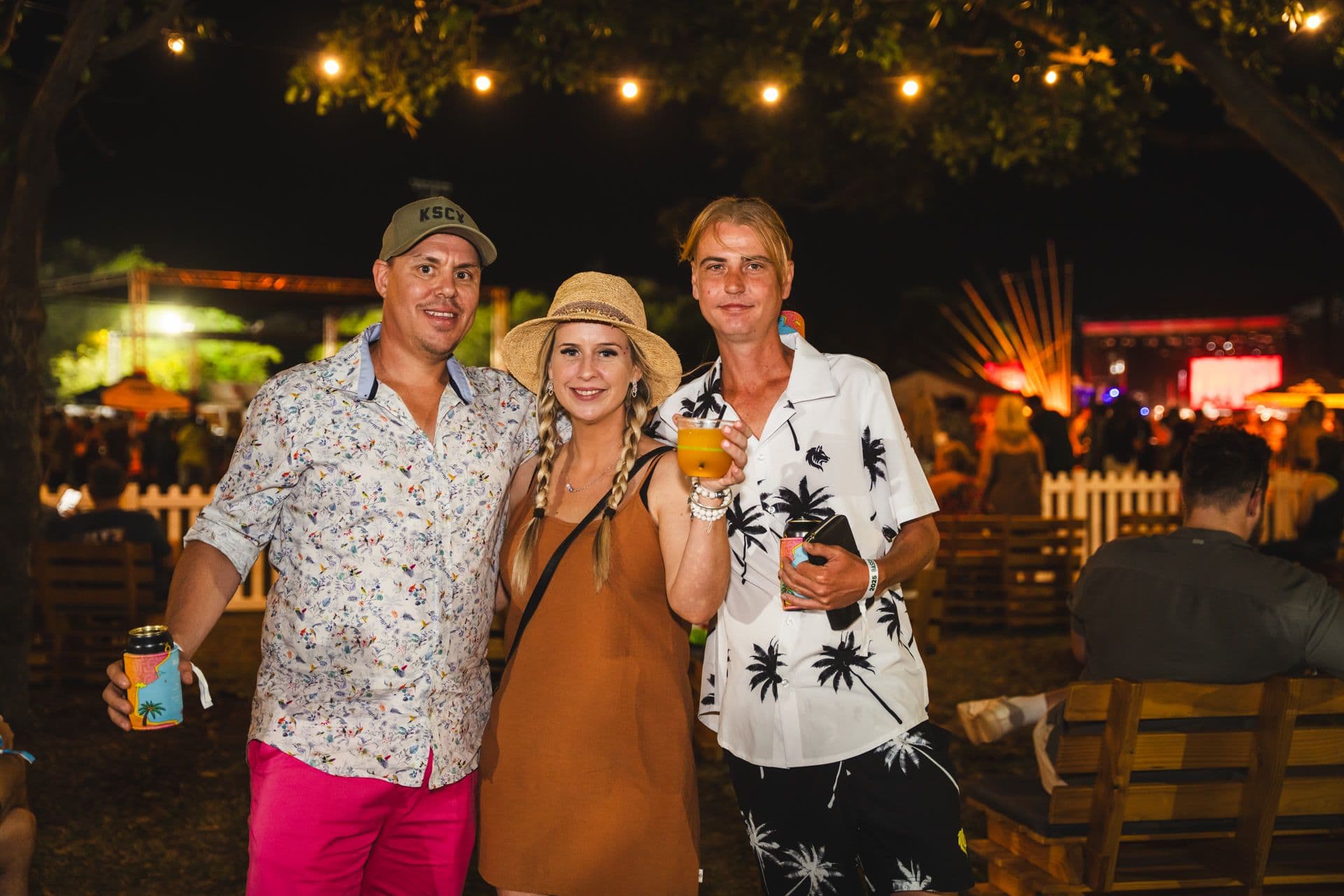 Three people smiling at an outdoor night event, with string lights and a stage in the background. They are holding drinks and dressed casually.