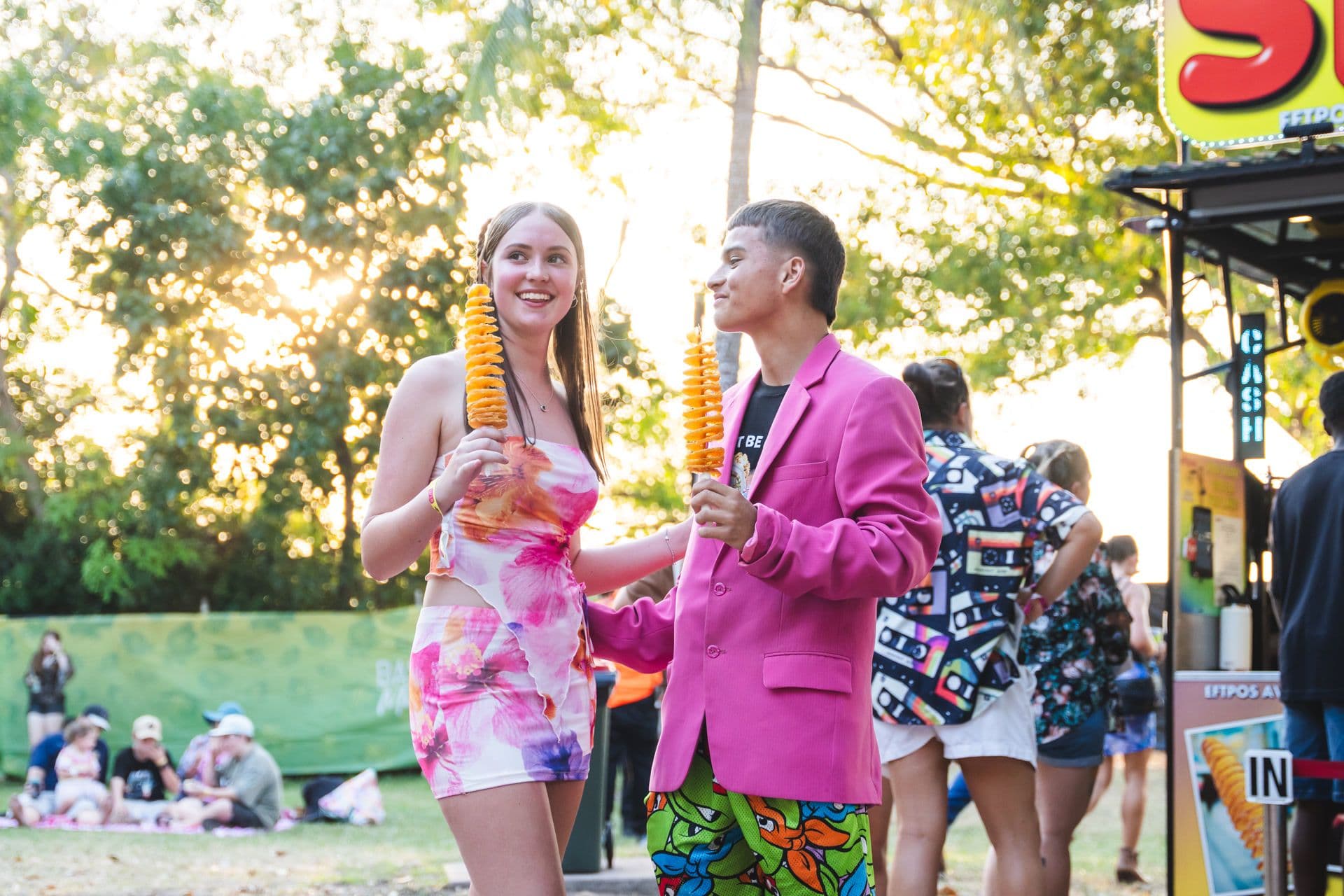 A smiling couple holds spiral-cut fried potatoes on sticks at an outdoor event, with people and trees in the background.