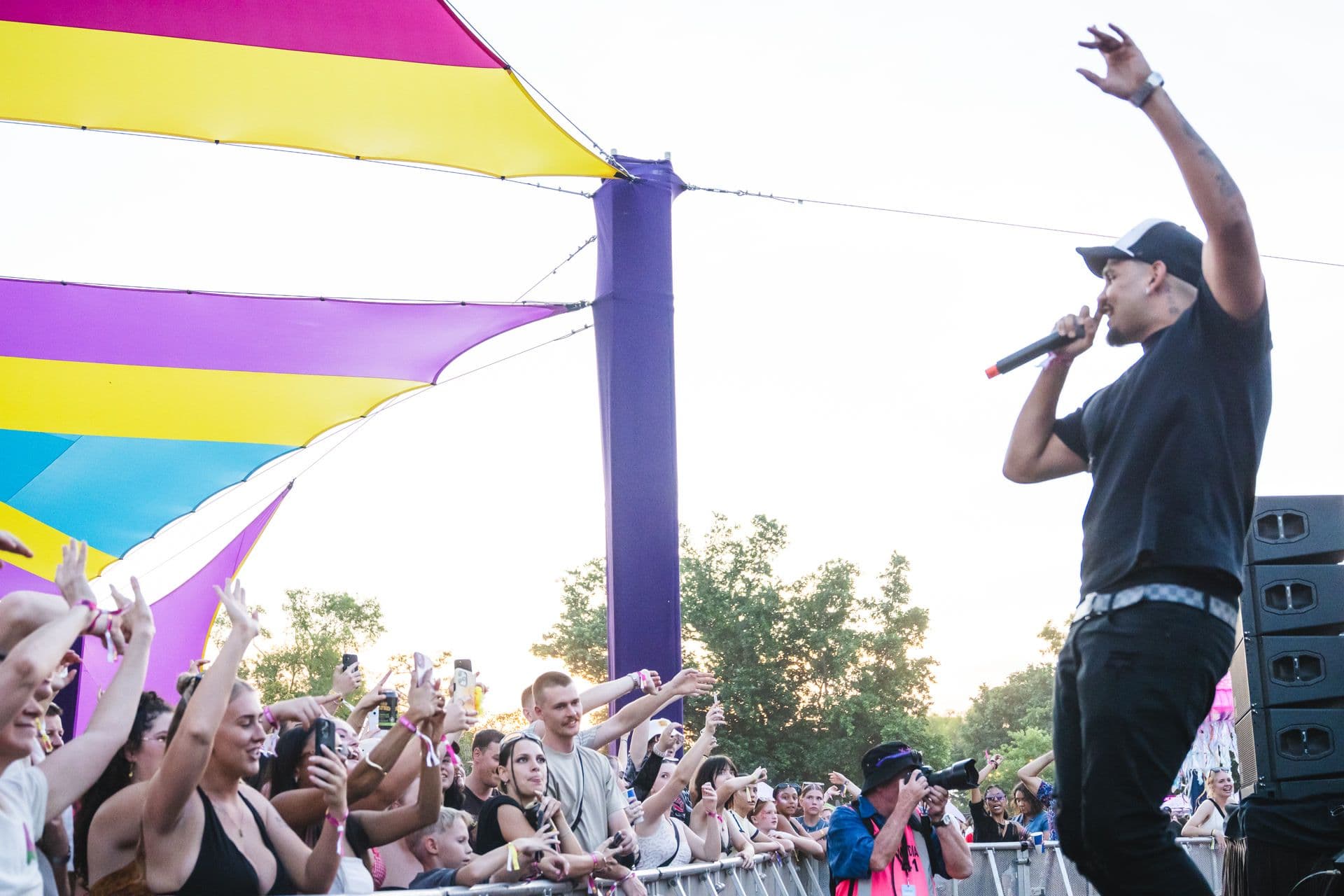 Performer on stage with a microphone, engaging a lively crowd at an outdoor event with colourful canopy overhead.