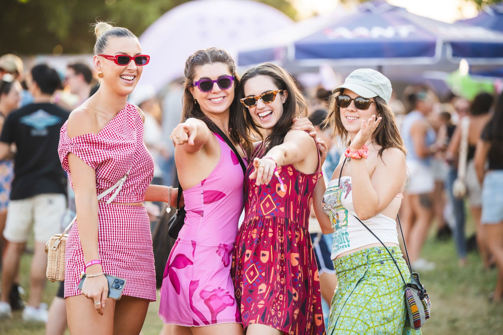 Four women in colorful outfits and sunglasses smile and point at the camera at an outdoor event, with a crowd and tents in the background.