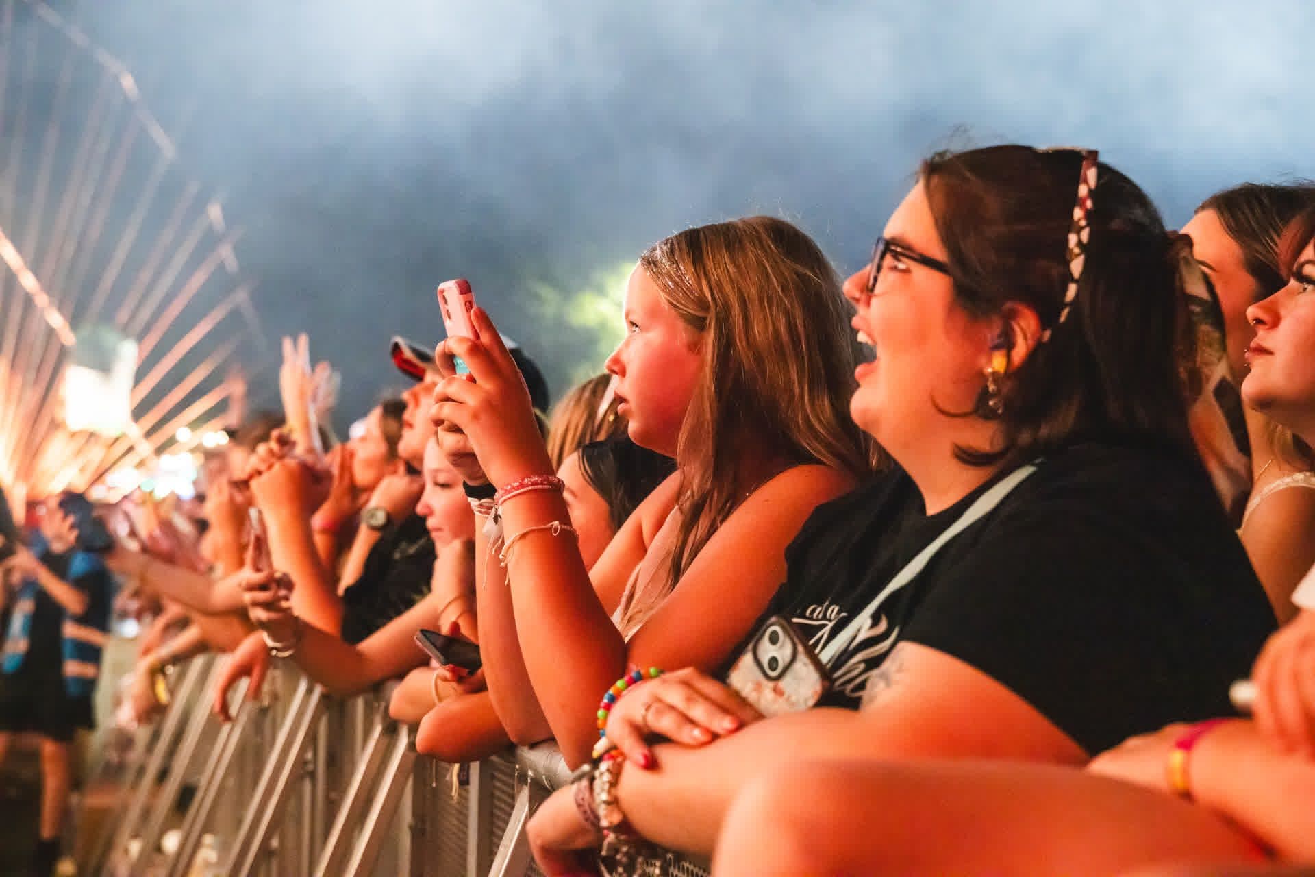 Audience at a concert, excitedly watching and recording with phones, with colorful lights and a ferris wheel in the background.