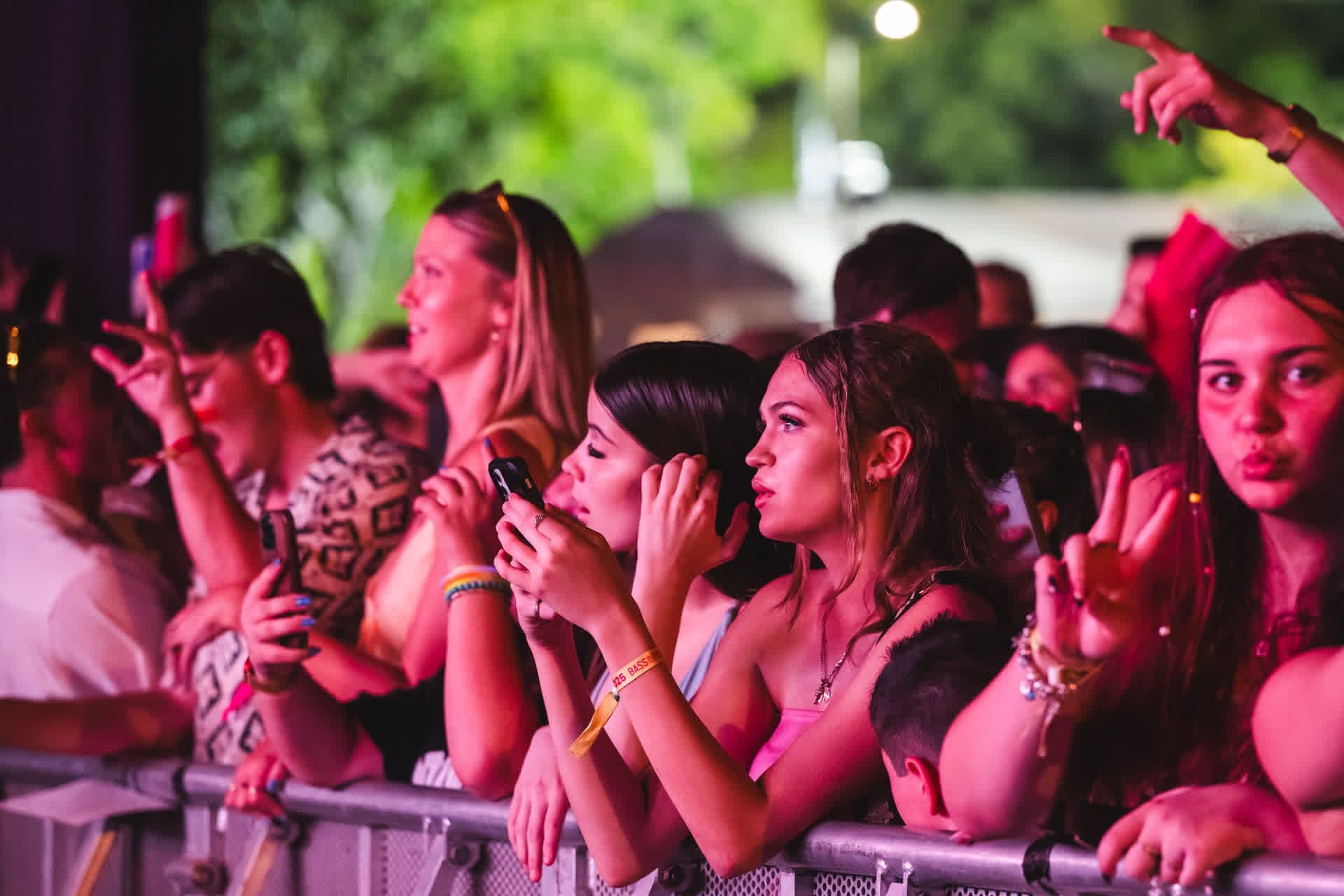 Audience at a concert, standing behind a barrier, some holding phones, with colorful lights and trees in the background.