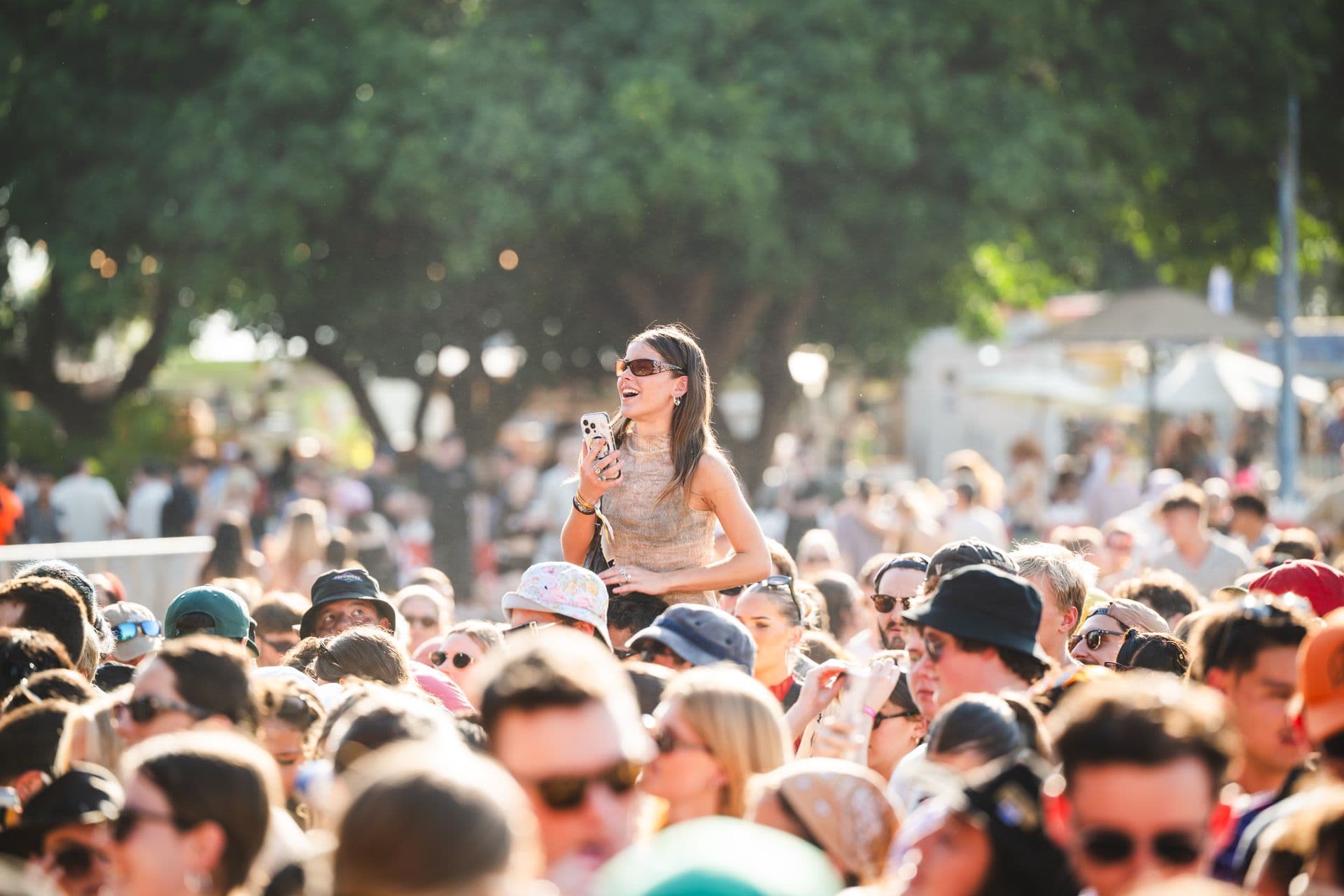 A woman stands above a large crowd at an outdoor event, holding a phone, with trees in the background.