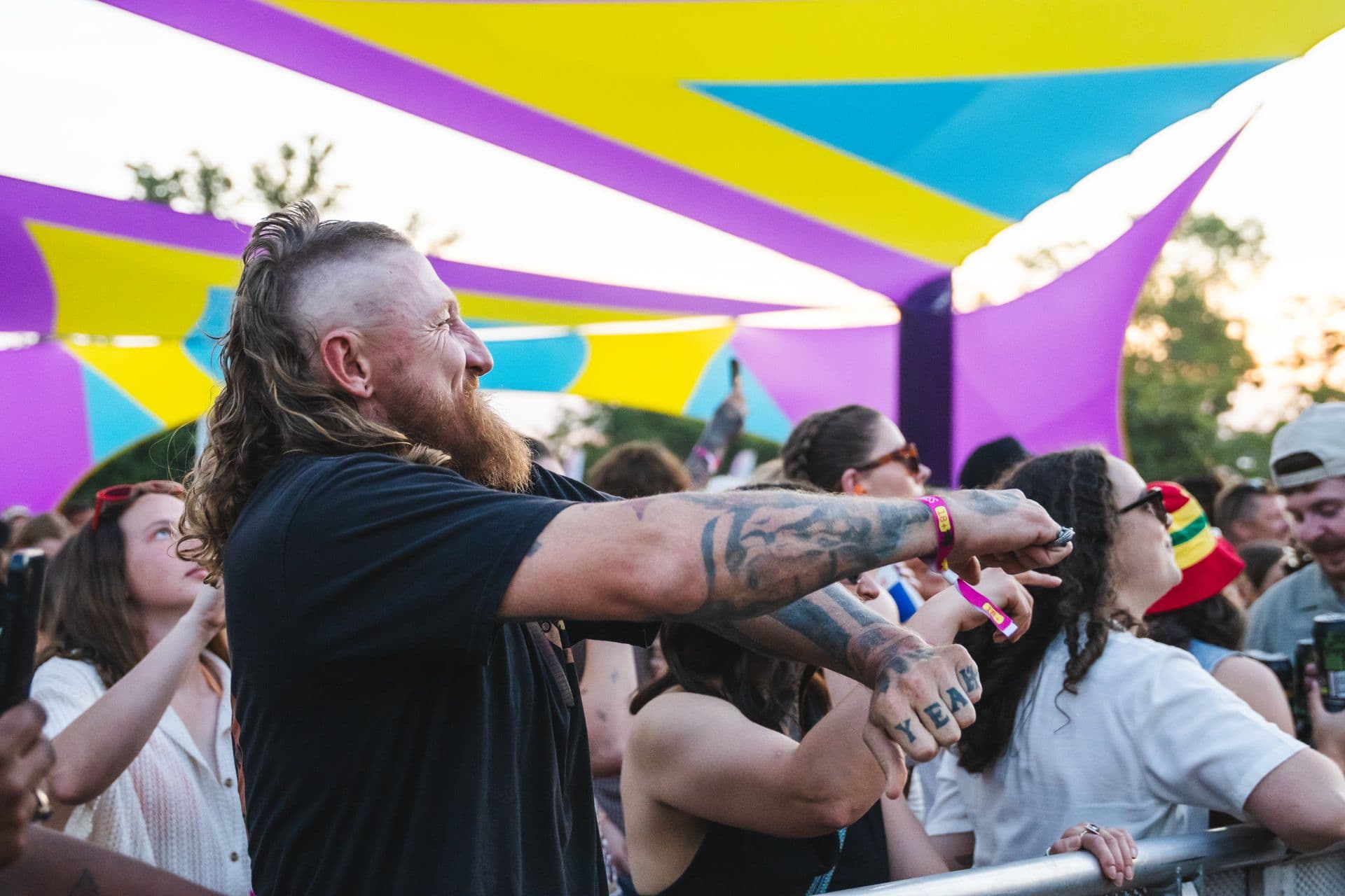 A bearded man with tattoos enjoys a lively outdoor event, surrounded by a diverse crowd under vibrant purple and yellow canopy tents.