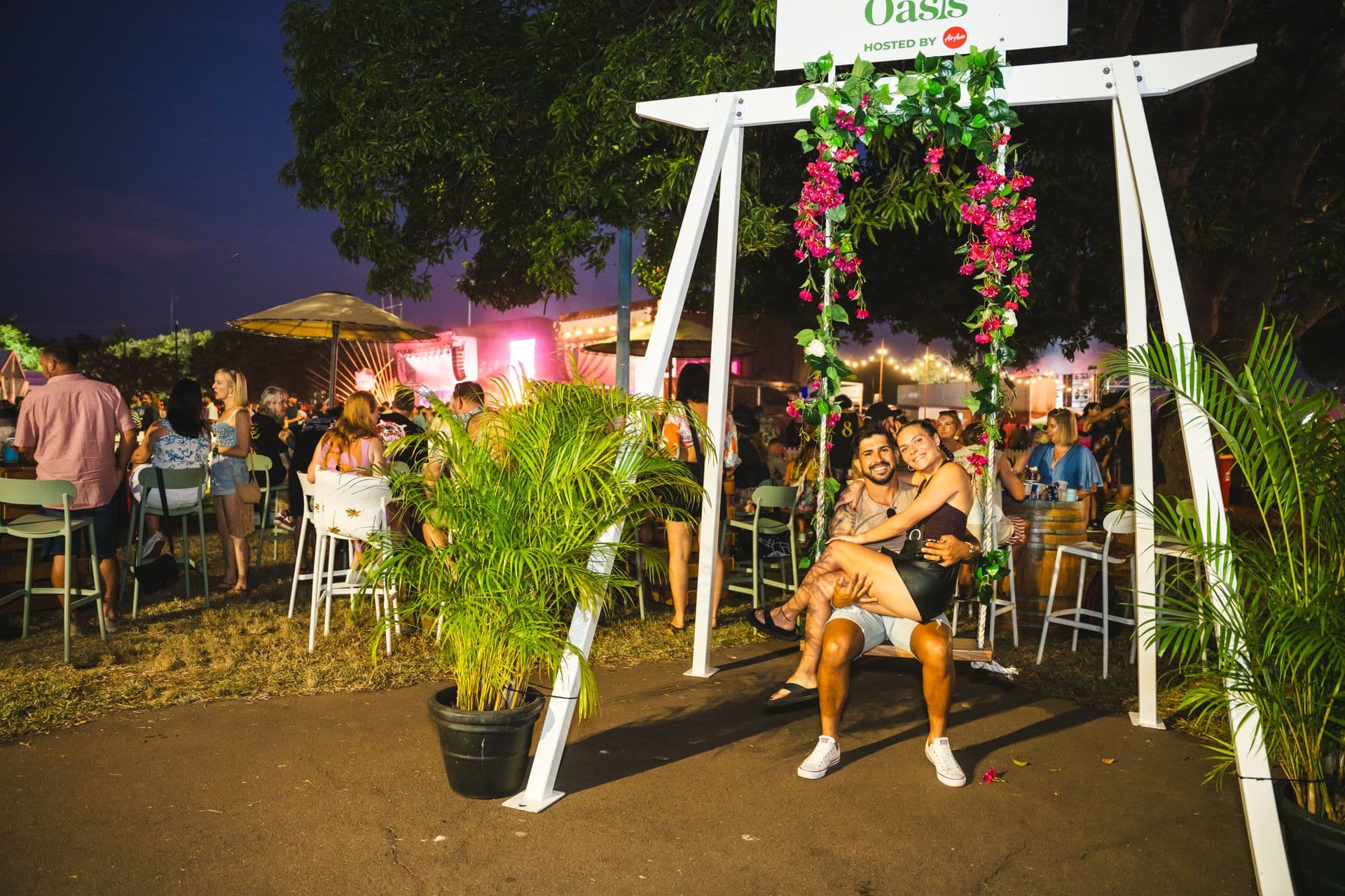 A couple sits on a decorated swing at an outdoor event, surrounded by people, plants, and lights in the evening.