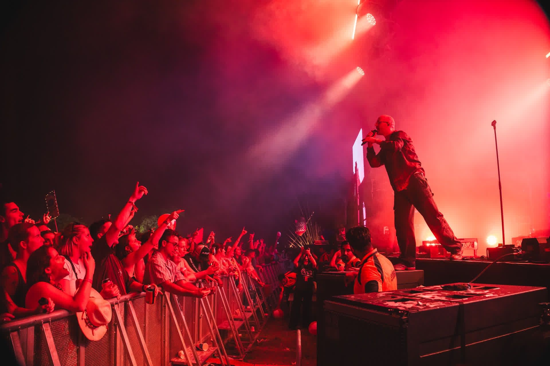 Performer on stage in red lighting, engaging with an enthusiastic crowd behind a barrier at a concert.