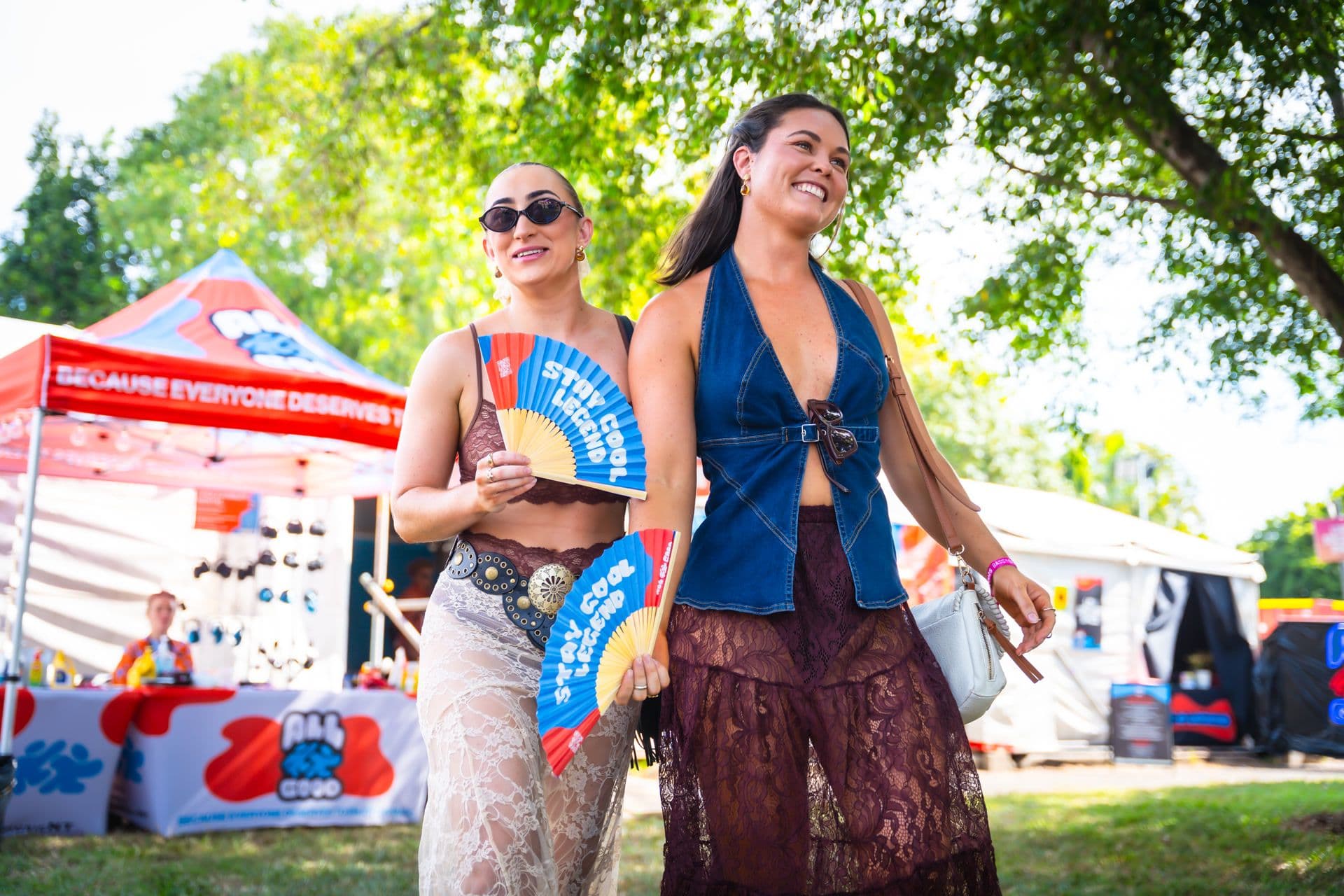 Two women smiling and walking at an outdoor event, holding colorful fans. Tents and trees are visible in the background.