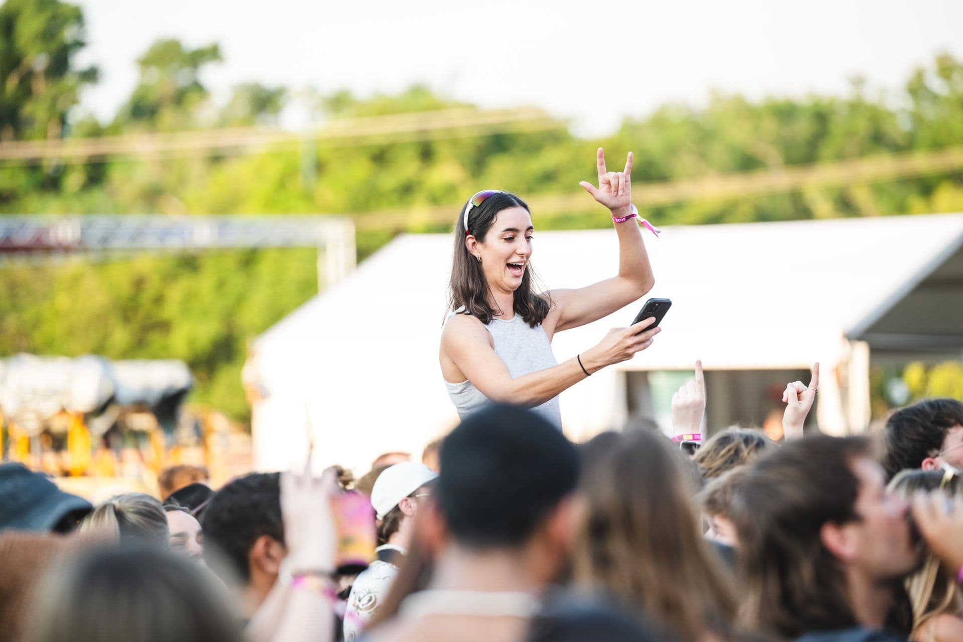 Woman on someone's shoulders at an outdoor concert, smiling, holding a phone, and making a hand gesture, surrounded by a crowd.