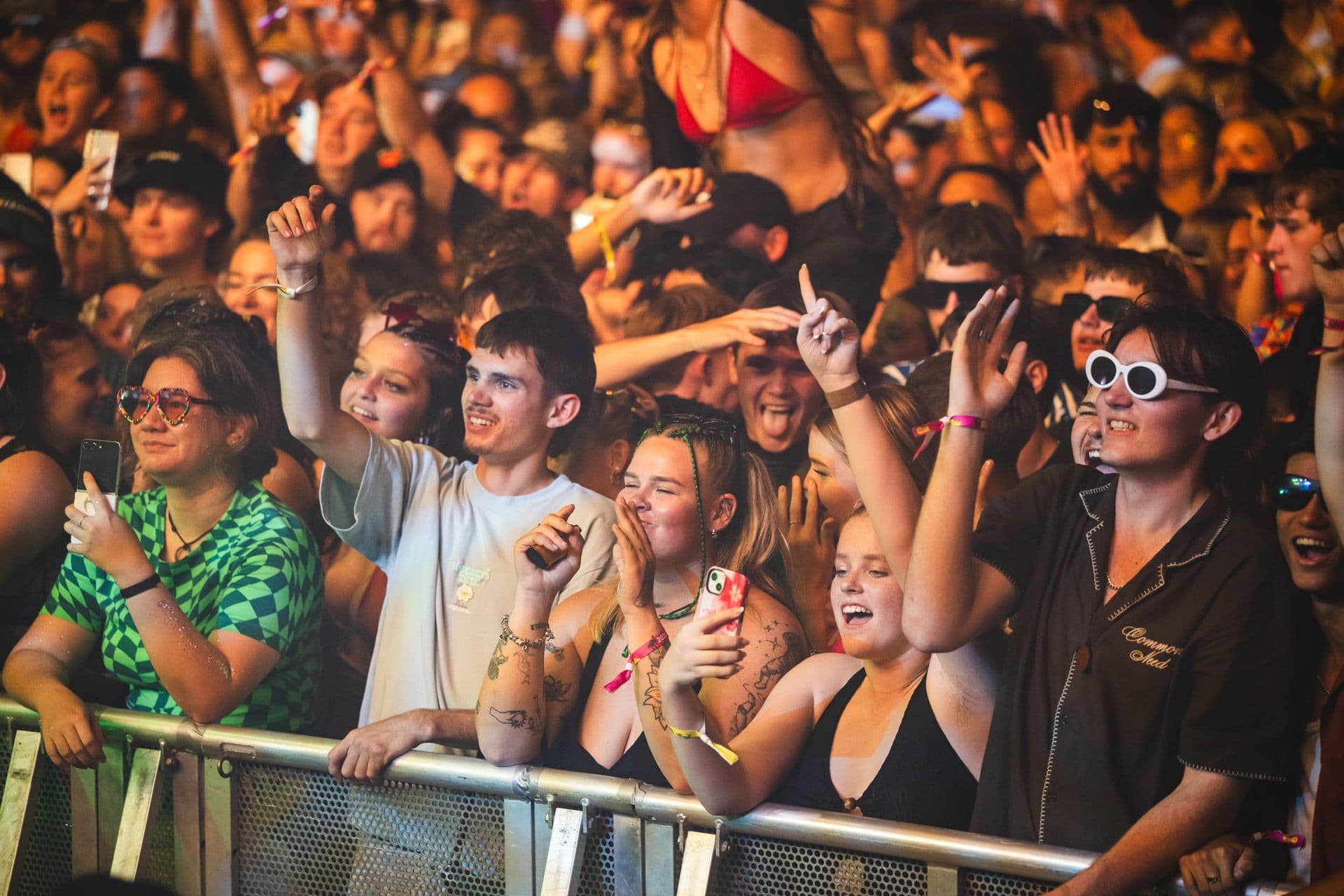 A lively crowd at a concert, with people smiling, cheering, and holding phones. Some wear sunglasses and colorful outfits, enjoying the event.