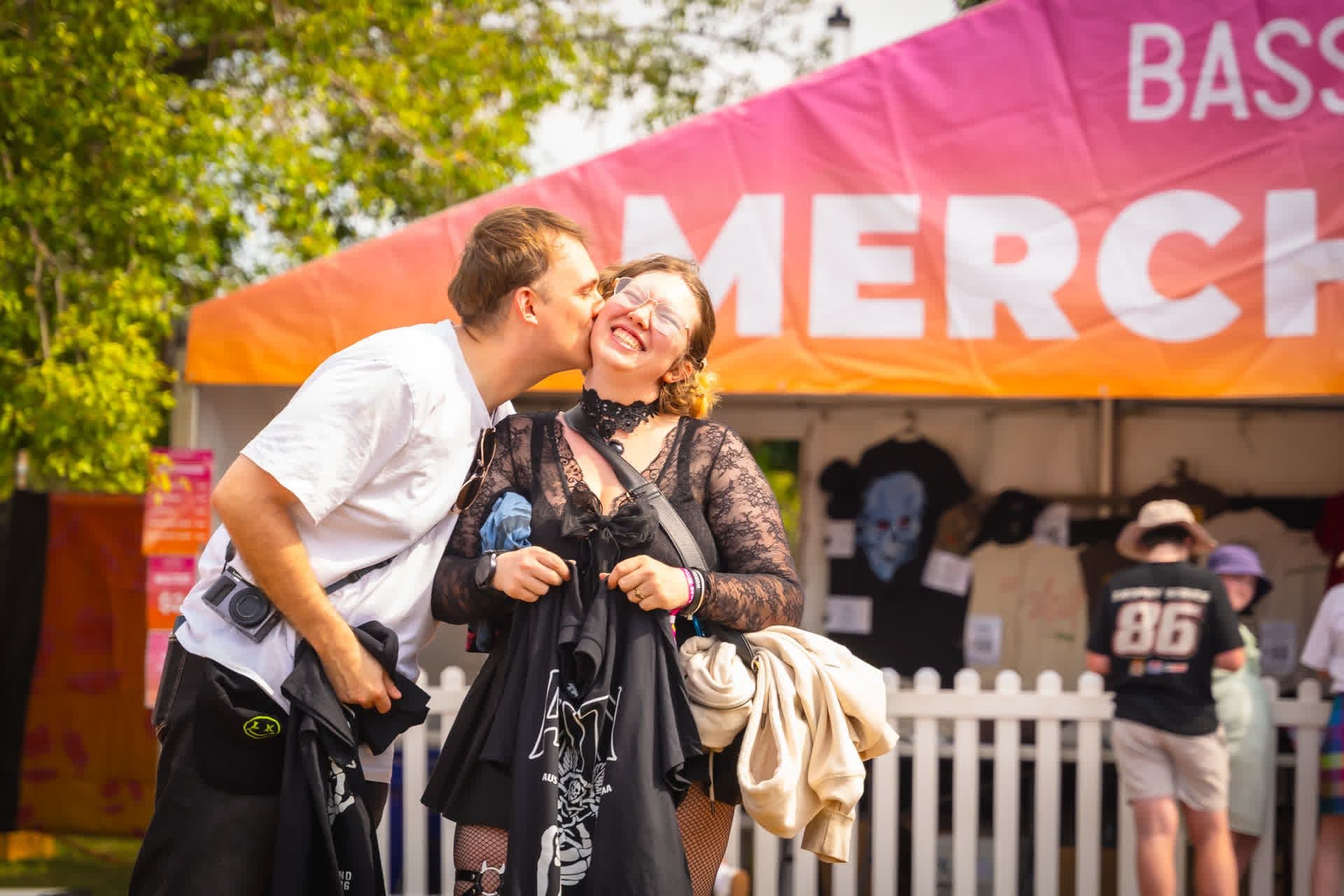 A person playfully kisses another on the cheek outside a merchandise tent at a festival. They hold T-shirts and smile, surrounded by a lively crowd.