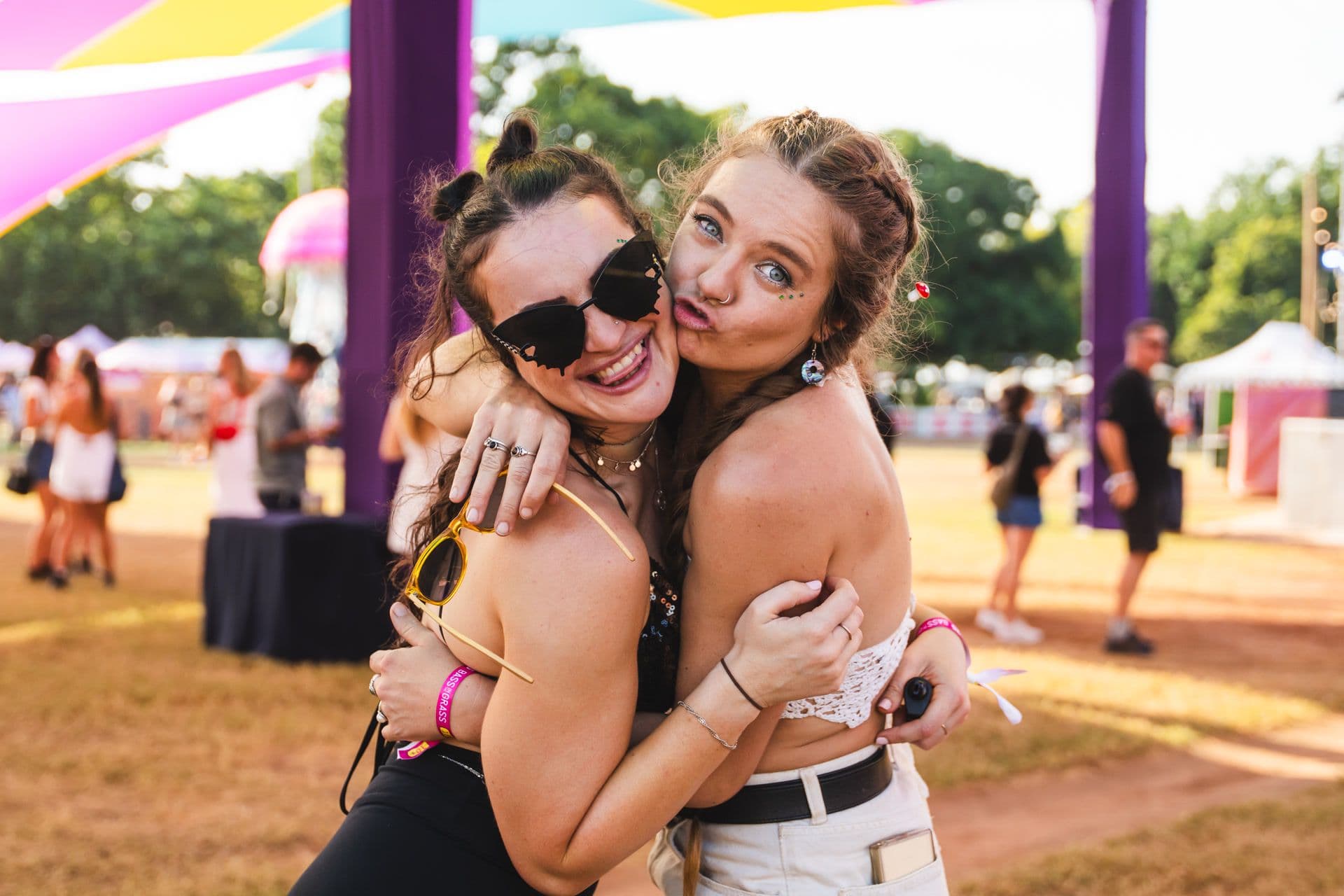 Two women smiling and hugging at an outdoor festival, wearing sunglasses and casual summer outfits, with colorful tents in the background.