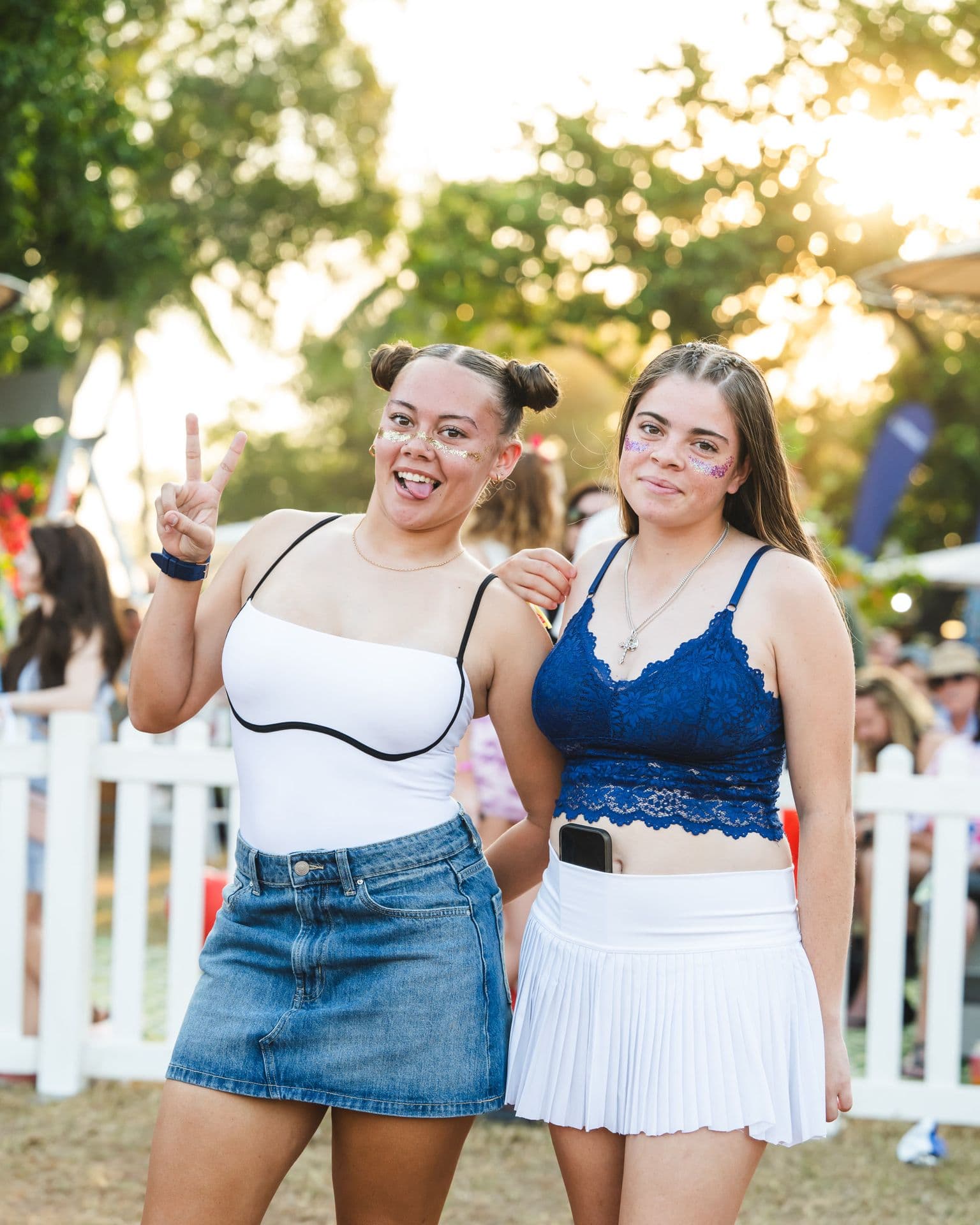 Two young women smiling outdoors, one making a peace sign. Both have glitter on their faces and are dressed in summer outfits.