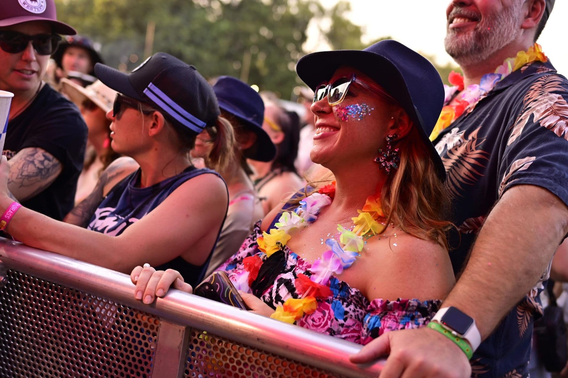 People at an outdoor event wearing hats and colorful leis, smiling and enjoying the atmosphere near a metal barrier.