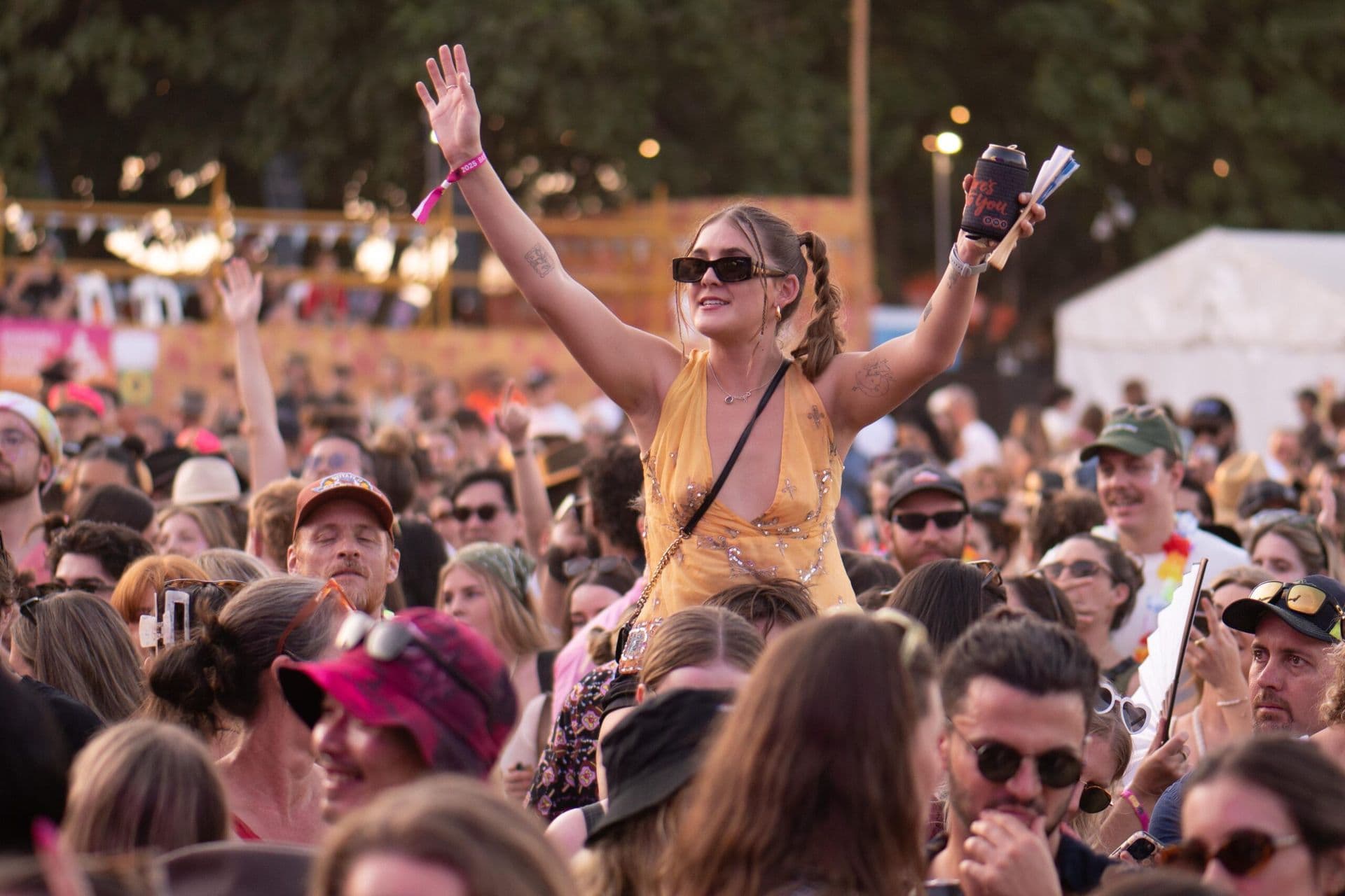A woman in a yellow dress, wearing sunglasses, is lifted above a festival crowd, smiling and raising her arms.