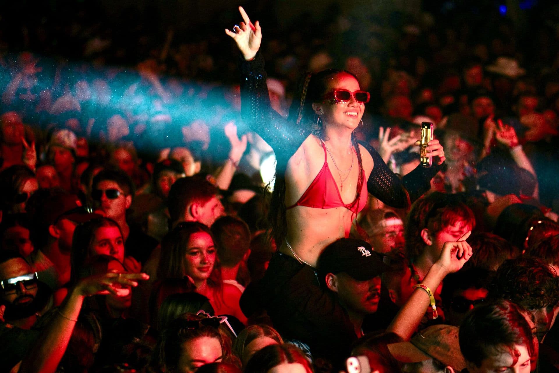 A woman in a red bikini top and sunglasses enjoys a concert, sitting on someone's shoulders amidst a lively crowd.