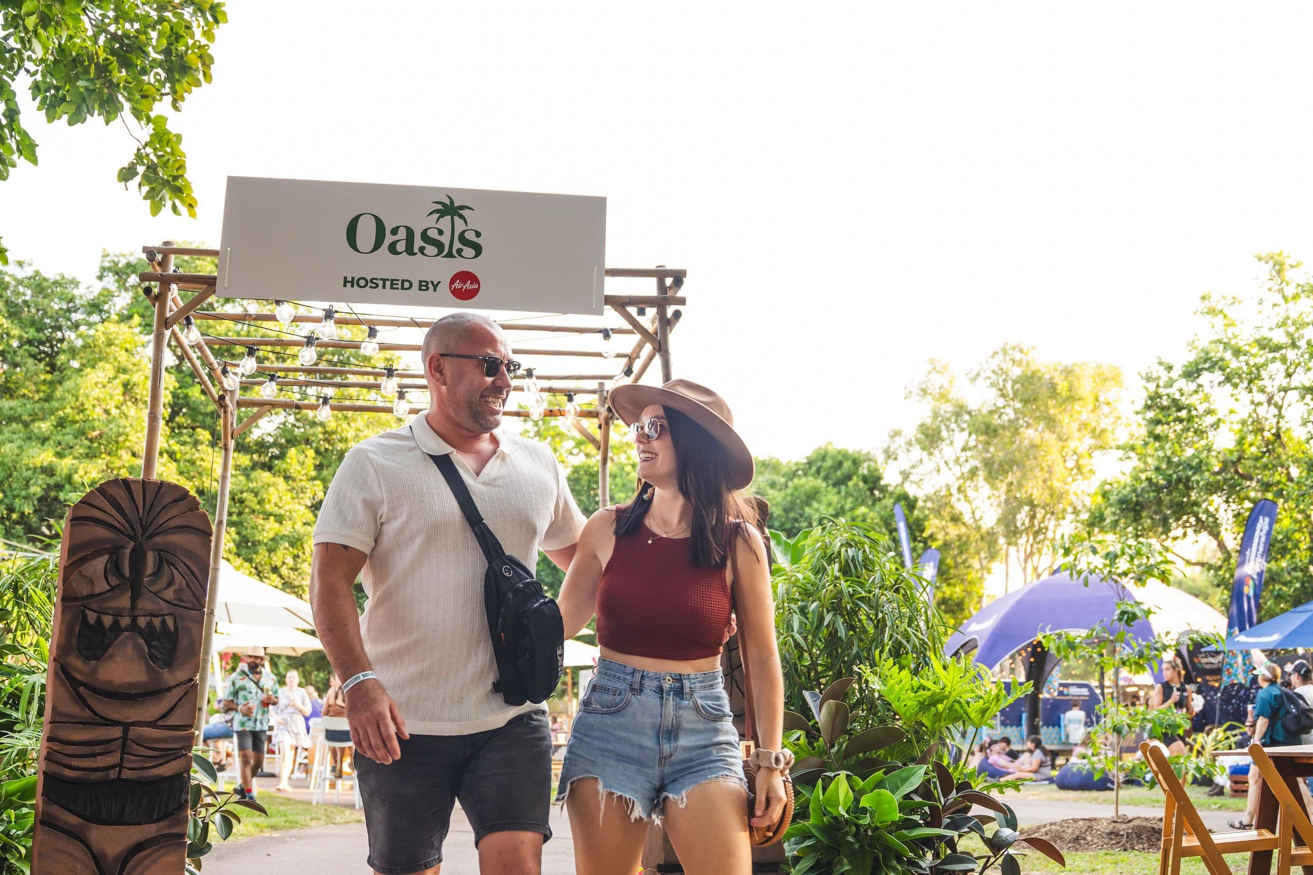 A couple walks through a tropical-themed outdoor event with lush greenery, a tiki statue, and a sign reading "Oasis."