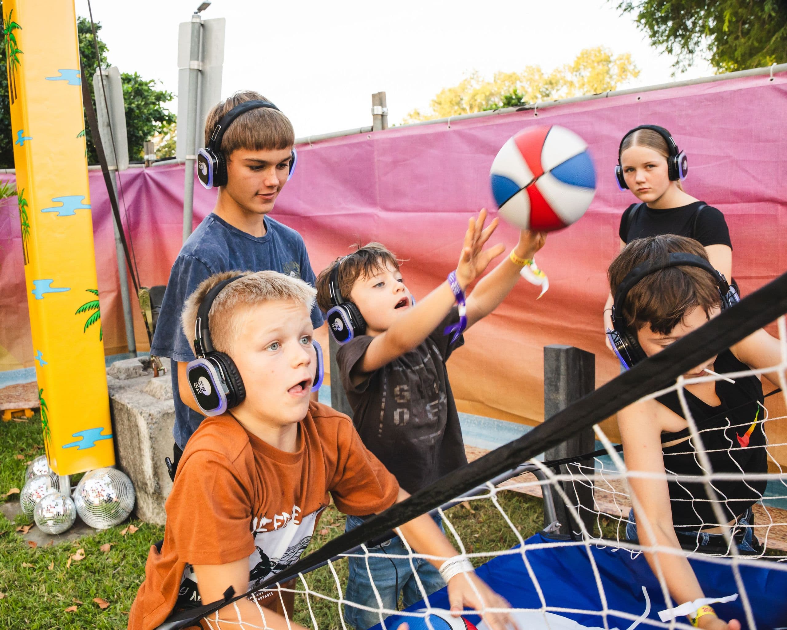 Children wearing headphones play an outdoor basketball game, enthusiastically reaching for the ball near a colorful backdrop.