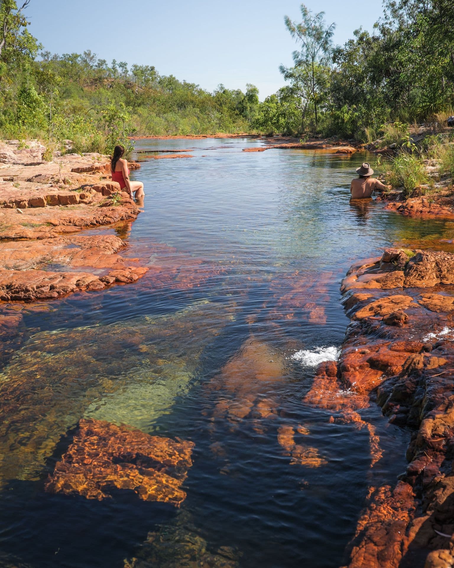 Two people relax by a clear, rocky river surrounded by lush greenery under a clear blue sky.