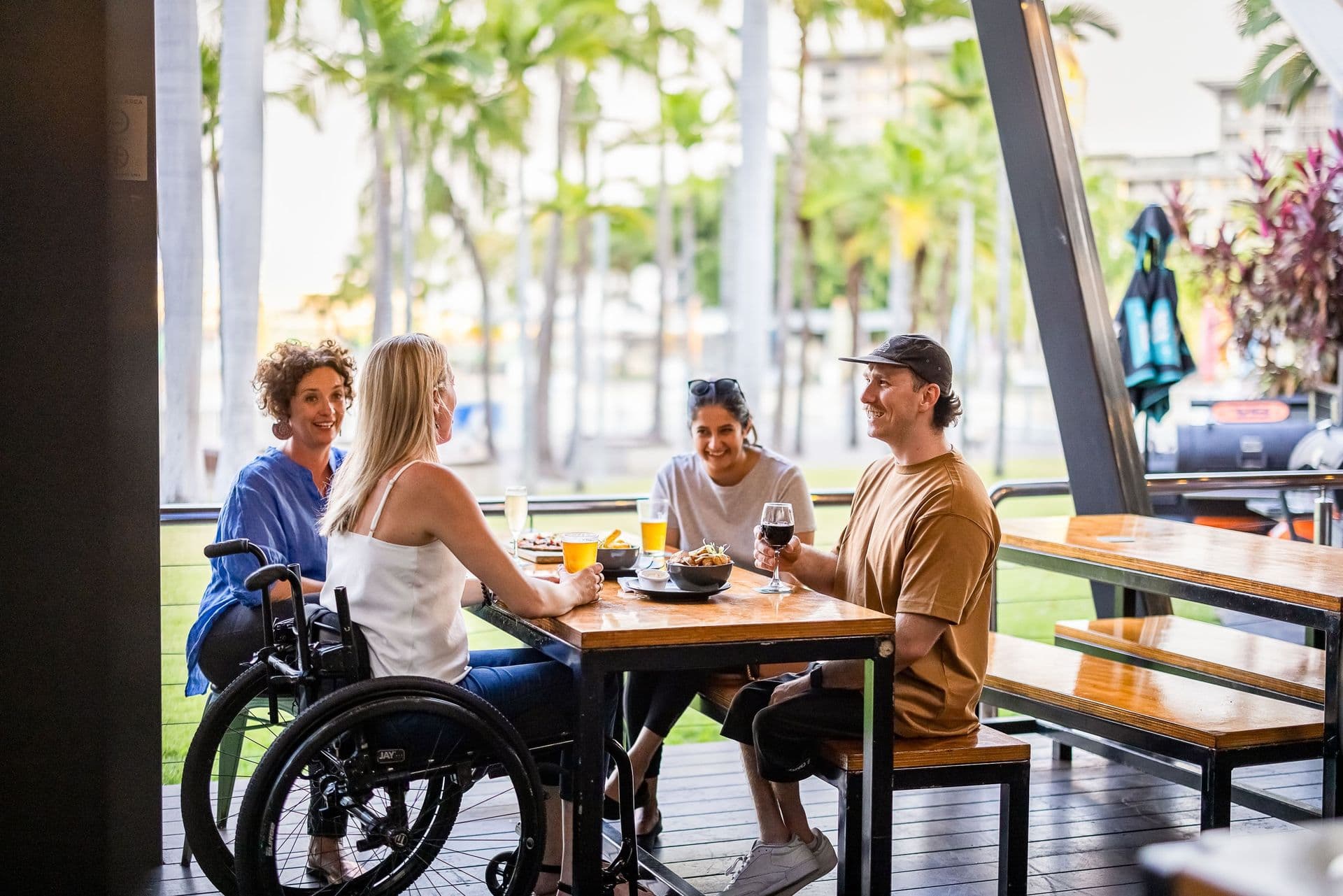 Four people sitting at an outdoor café table, chatting and enjoying drinks. One person is in a wheelchair. Palm trees are in the background.