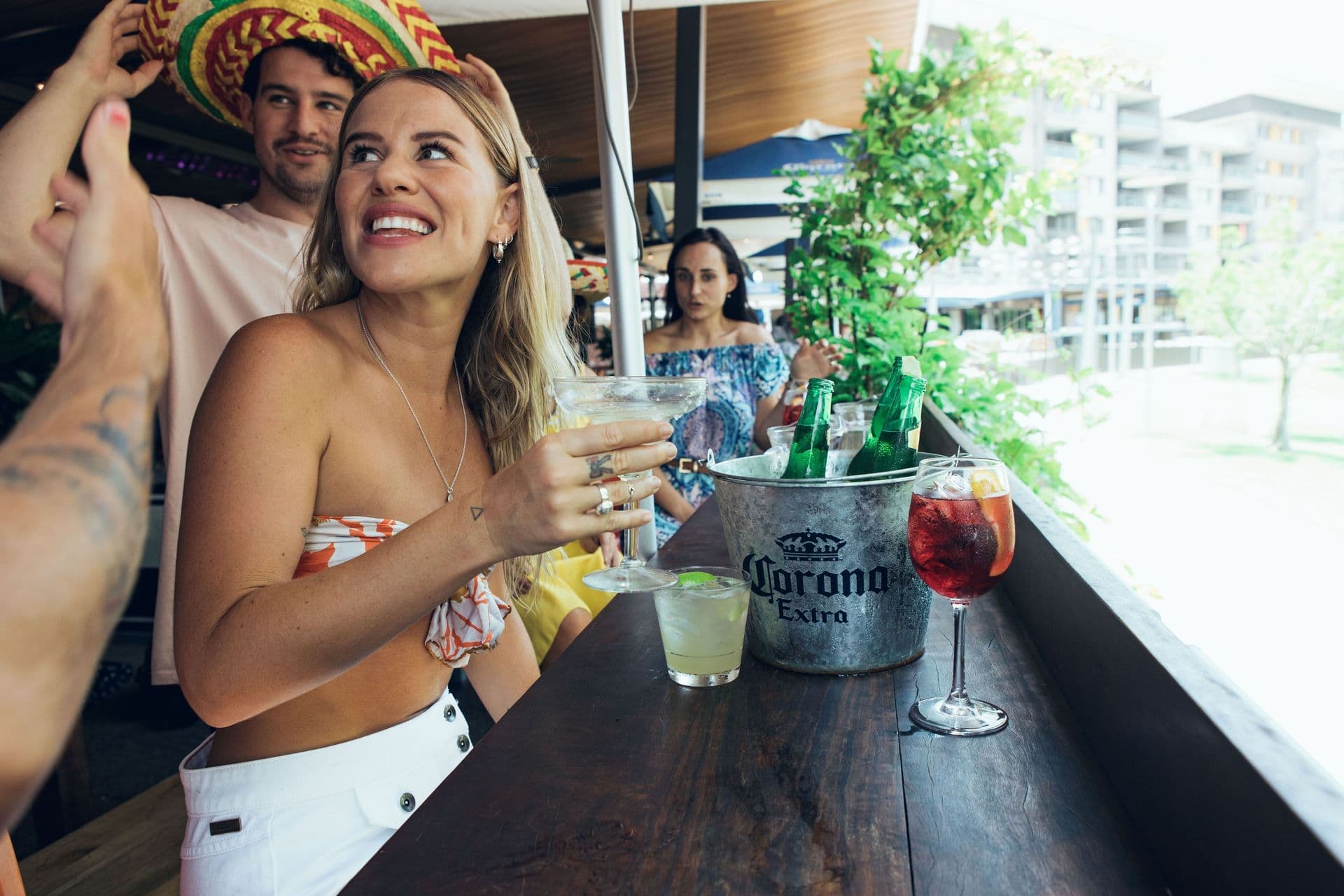 Woman smiling, holding a margarita at an outdoor bar. Ice bucket with drinks and people in festive attire in the background.