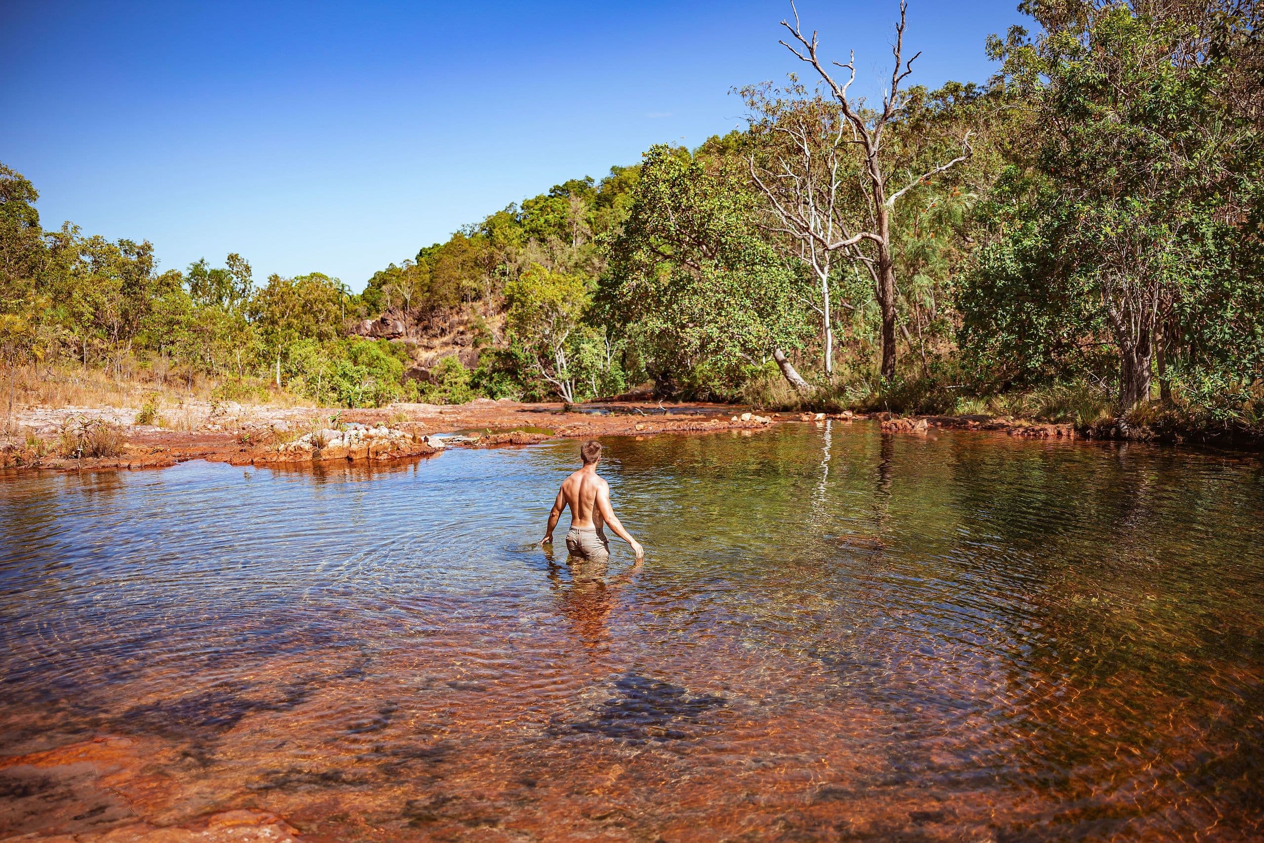 Person wading in a clear, shallow river surrounded by lush greenery and a bright blue sky.