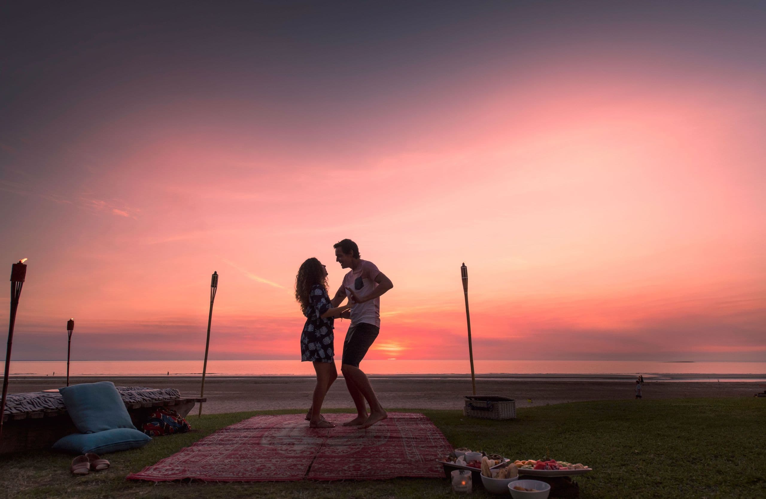 A couple dances on a beach at sunset, surrounded by torches, cushions, and a picnic setup. The sky is a vibrant blend of orange and pink hues.