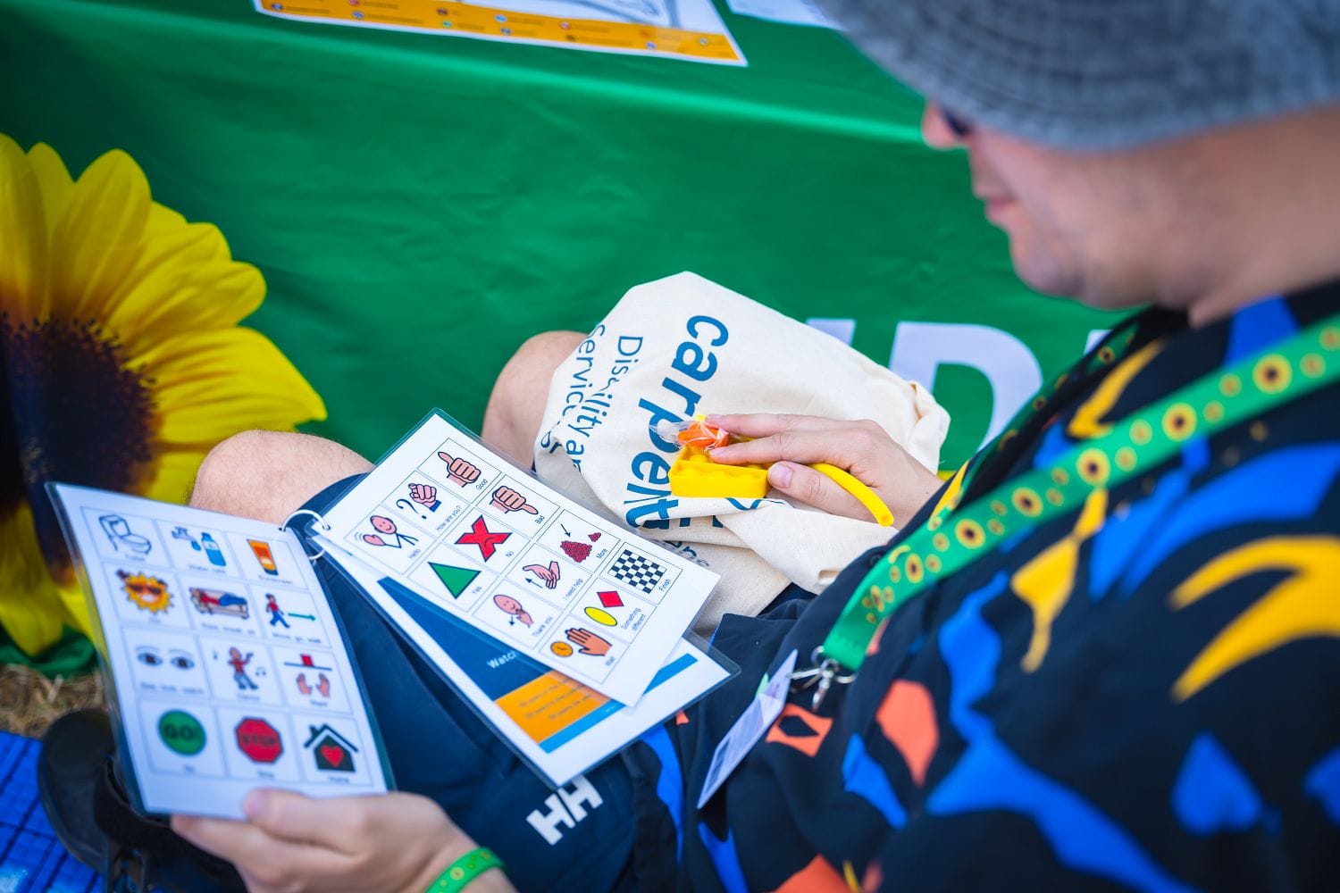 A person in a colorful shirt and hat examines pictorial cards on a mat. A sunflower and green background are partially visible.