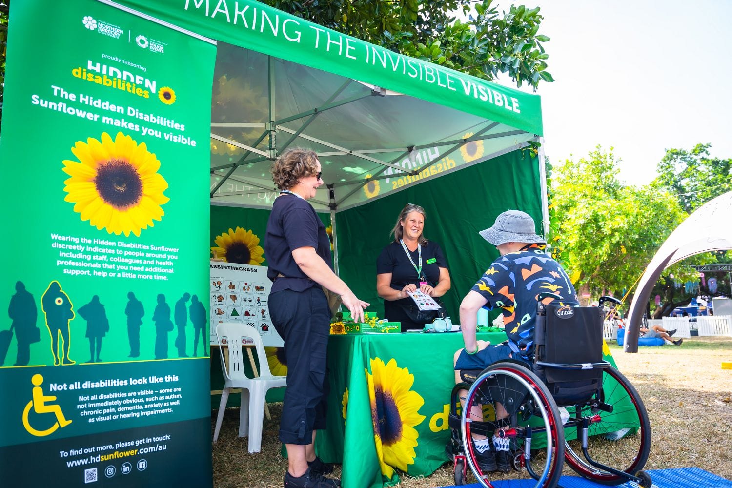 Two people at a green booth with "Hidden Disabilities Sunflower" branding, interacting with a person in a wheelchair.