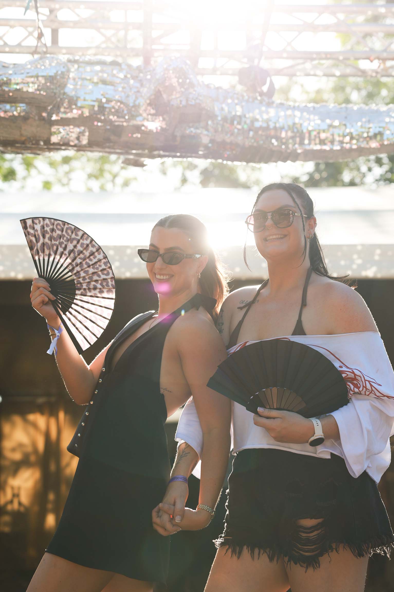 Two smiling women, wearing sunglasses, hold hands and fans outdoors under bright sunlight. One fan is leopard print, the other black.