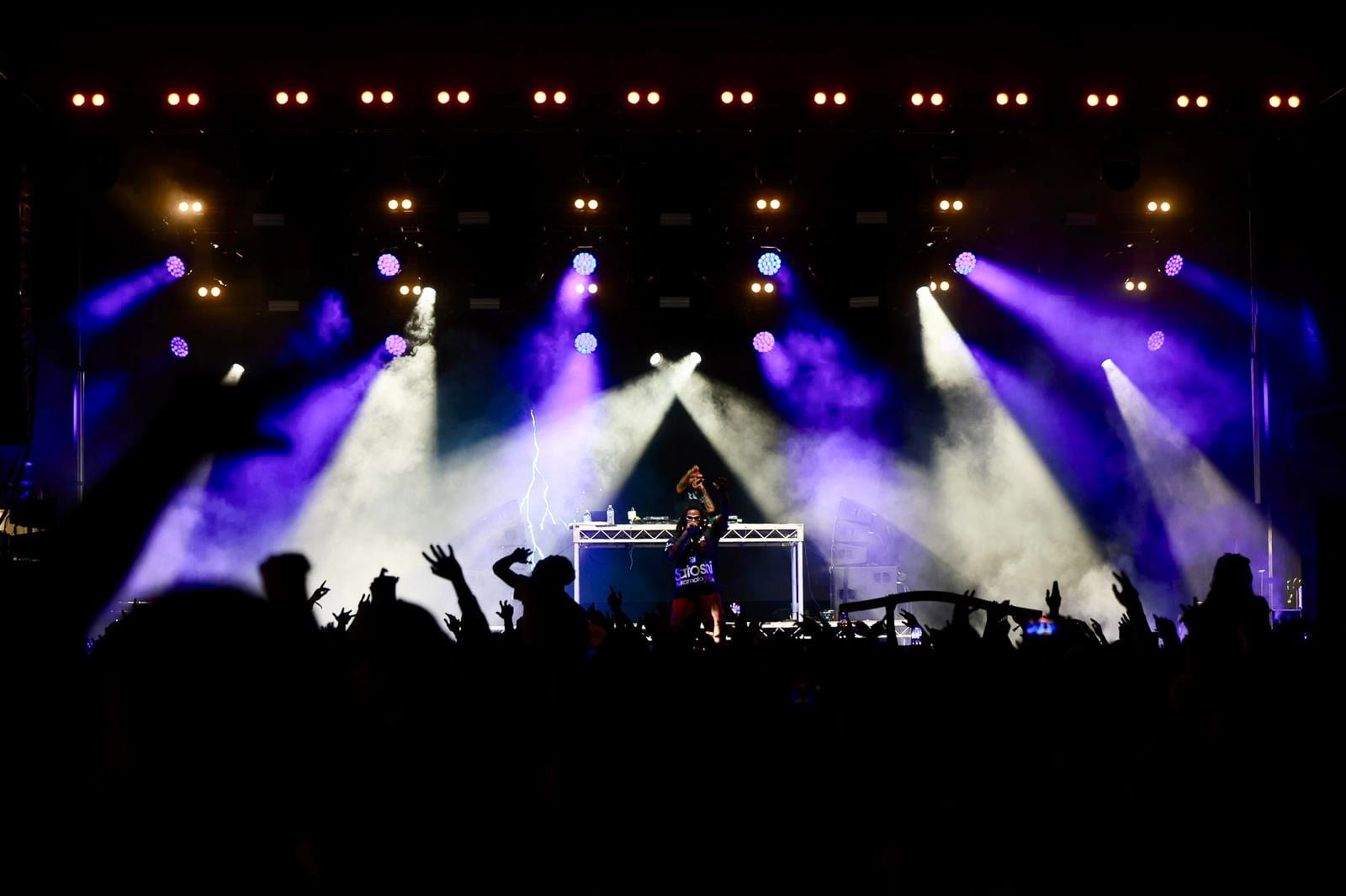 Nighttime concert with colorful stage lights and beams, a DJ at the mixer, and a silhouetted cheering crowd.