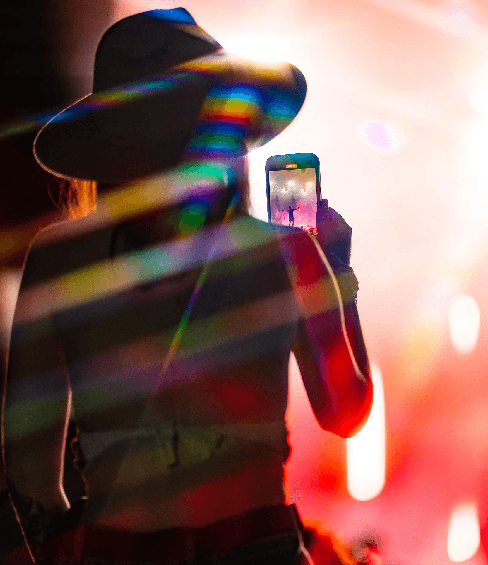 A person in a hat takes a photo with a smartphone at a concert, with colorful lights and lens flare in the background.
