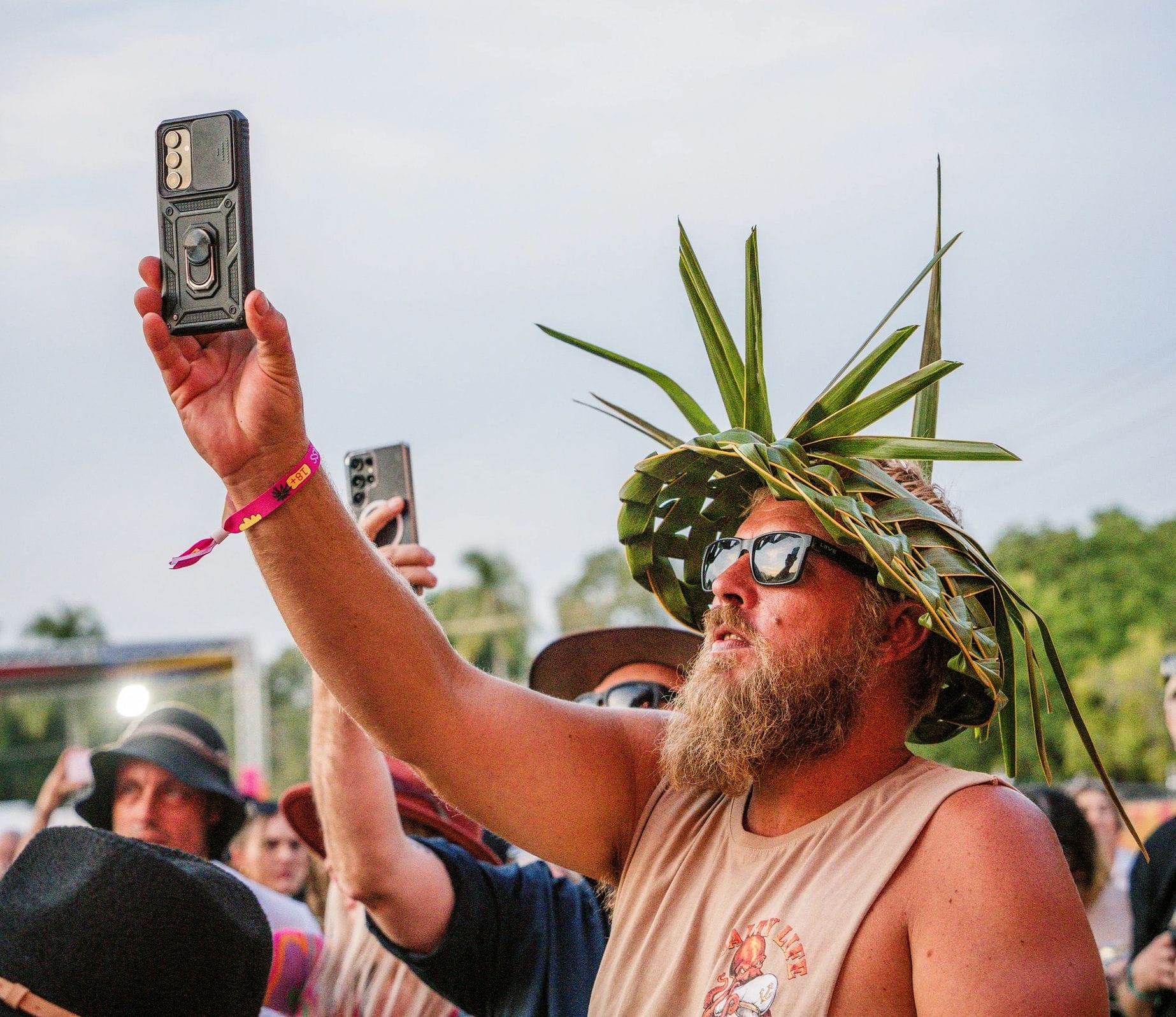 Man with a beard wearing a palm leaf hat and sunglasses, holding up a phone at an outdoor event, surrounded by a crowd.