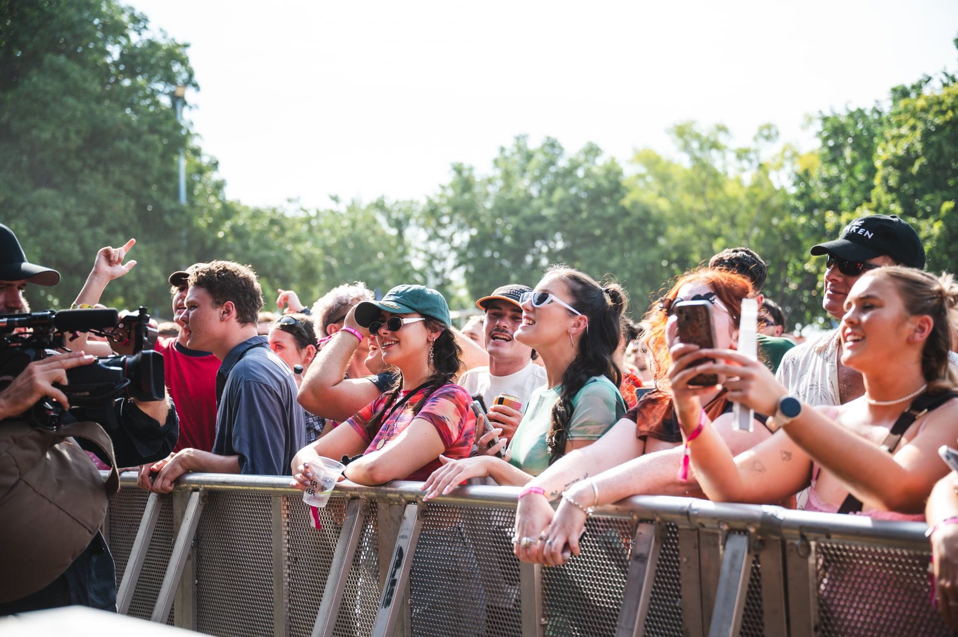 A lively crowd at an outdoor concert, smiling and cheering near the stage, with trees in the background on a sunny day.