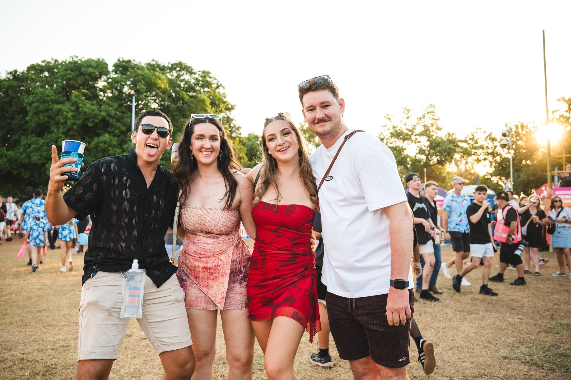 Four people smiling and posing together at an outdoor festival, surrounded by a crowd and trees in the background.