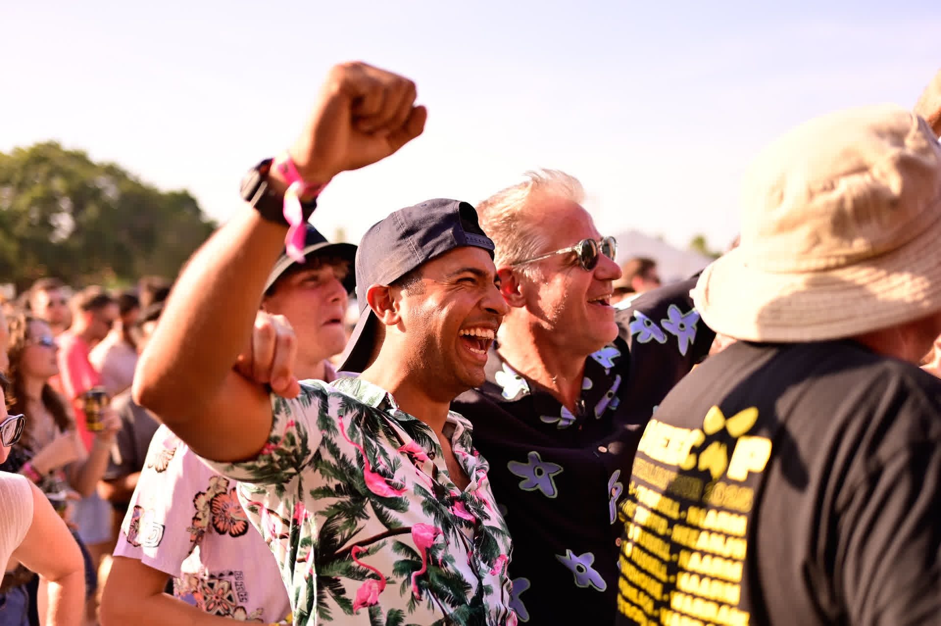 A crowd of people enjoying an outdoor event, with two men in floral shirts smiling and cheering. Sunny weather and clear skies in the background.