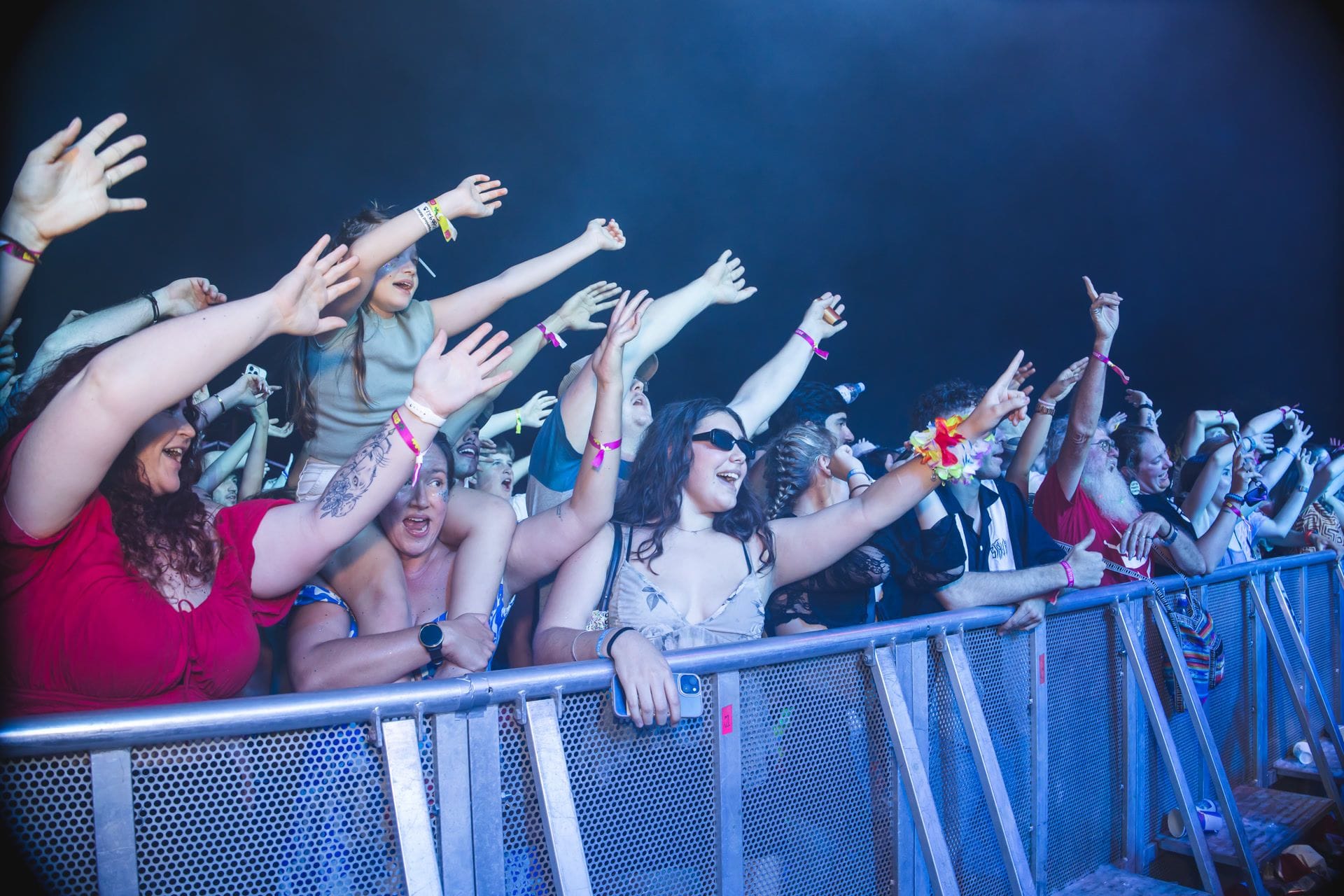 A lively crowd at a concert, with people raising their hands and cheering near a barrier under vibrant stage lighting.