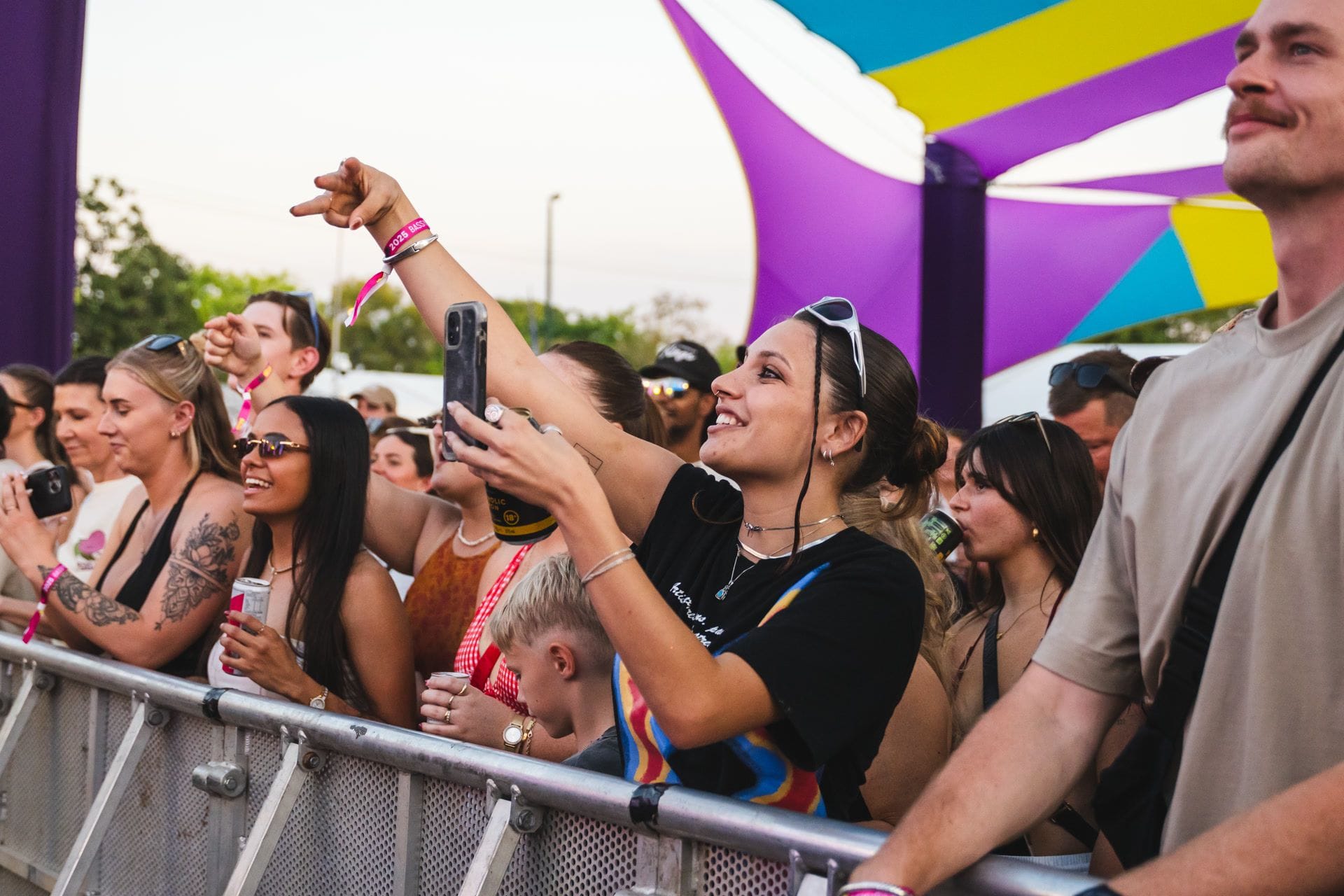 People enjoying a lively outdoor concert, some holding phones to capture the moment, under colourful canopy structures.
