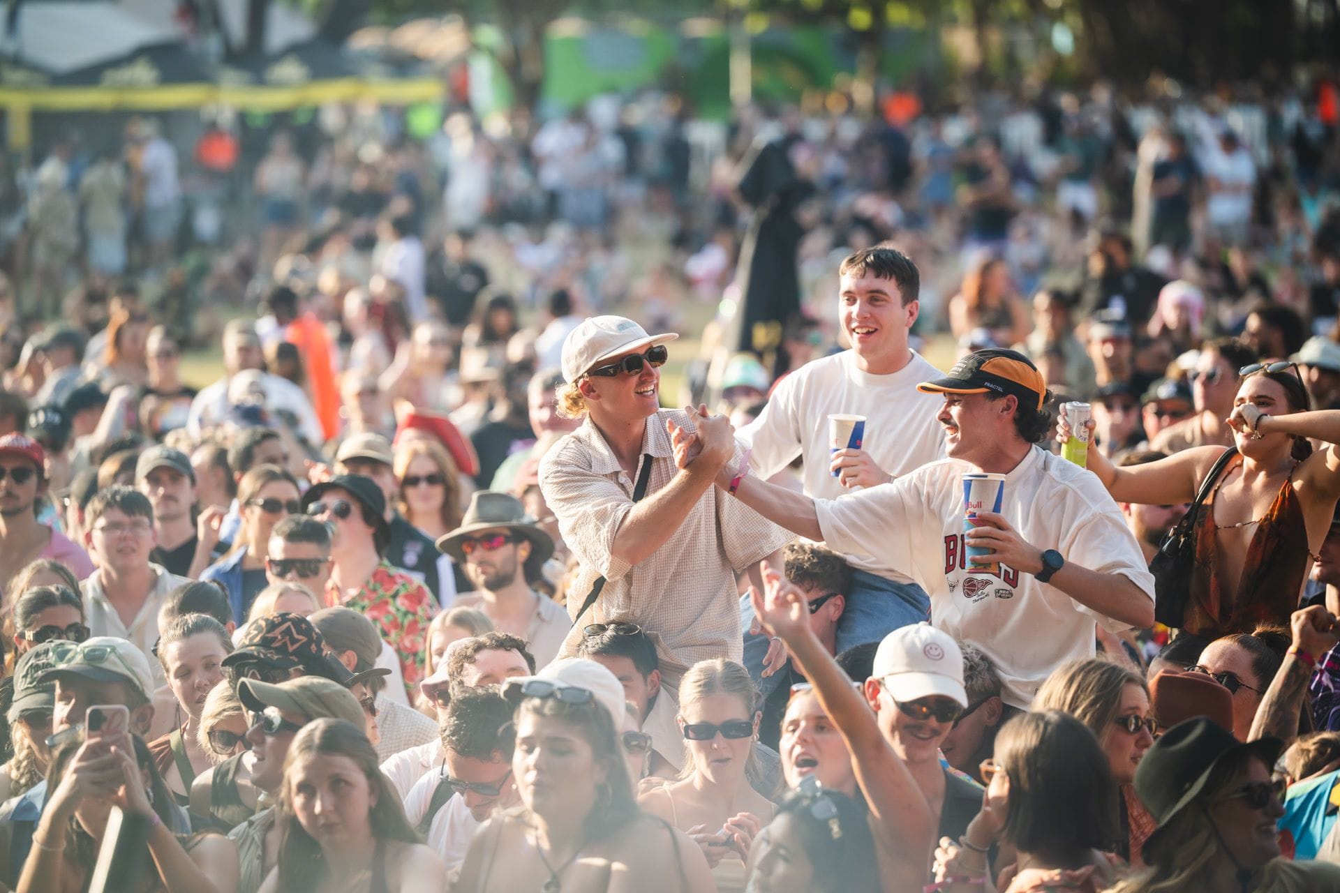 A lively outdoor crowd enjoys a music festival, with people cheering, wearing sunglasses, and holding drinks under a sunny sky.